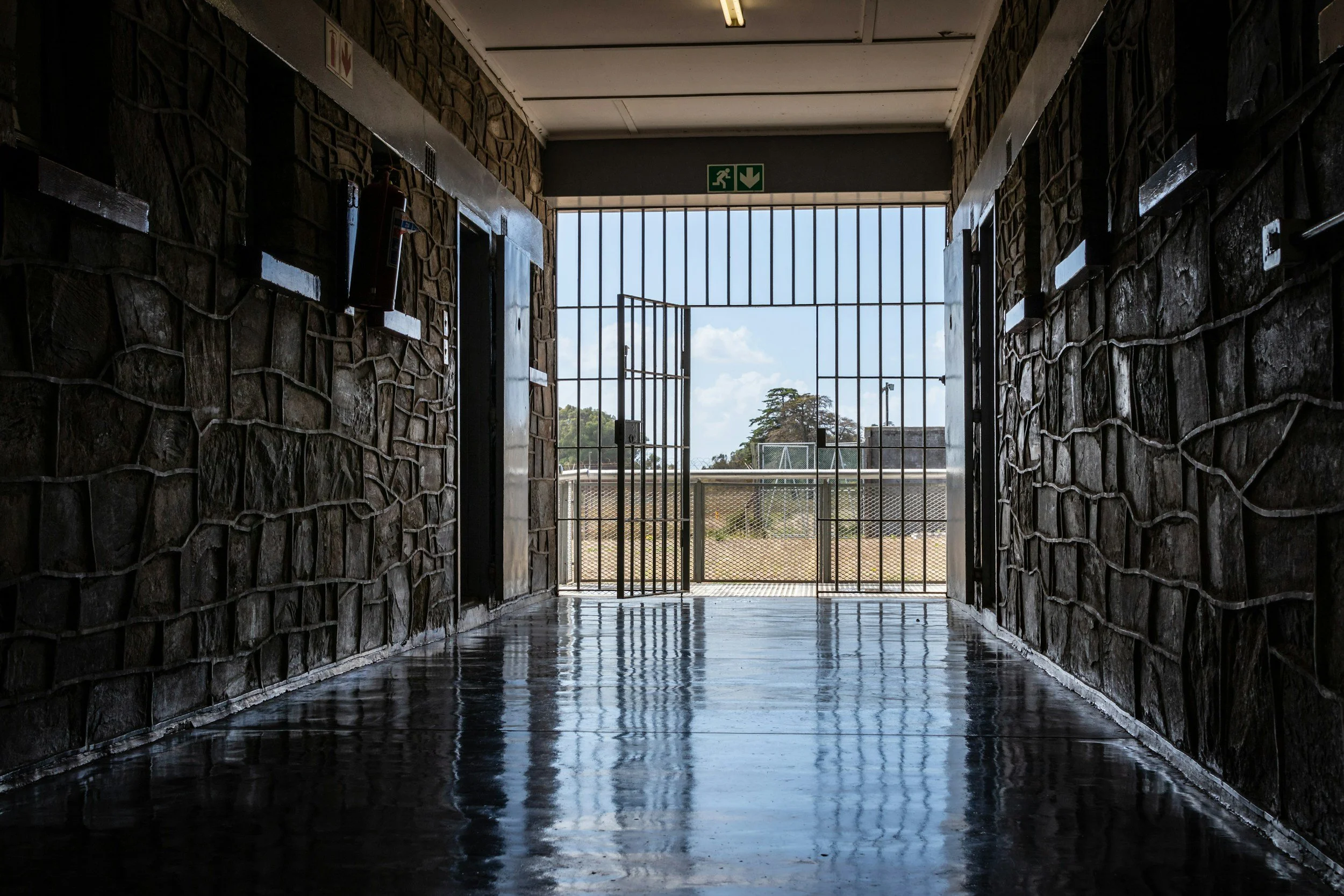 A covered outdoor pathway with stone walls, a polished floor, and a metal grille gate leading to an open space with a sports field and trees under a blue sky.