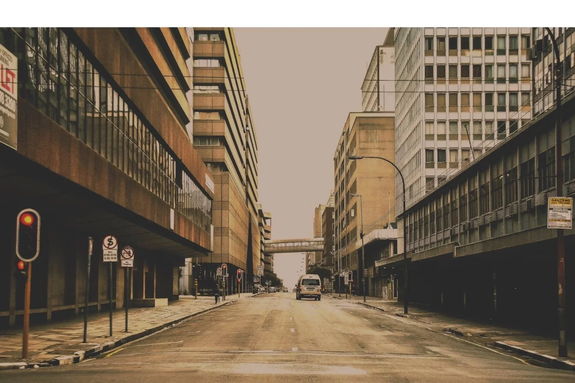 Deserted city street with tall office buildings, traffic lights, and a white van in the distance during daytime.