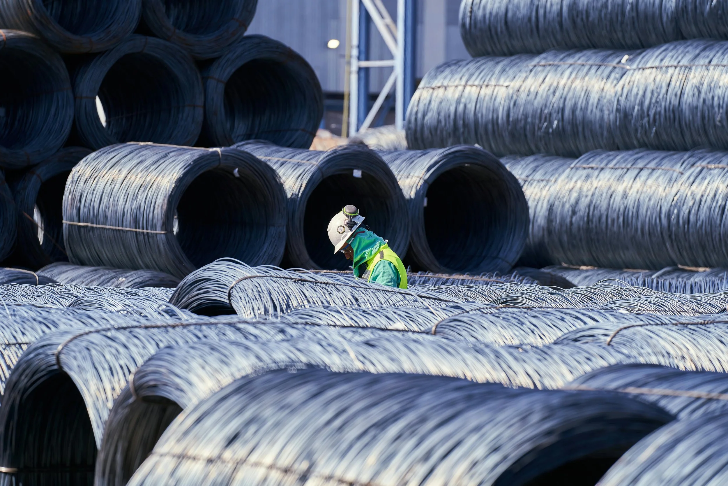 A construction worker wearing a helmet and safety gear walking among large steel pipes stacked outdoors.