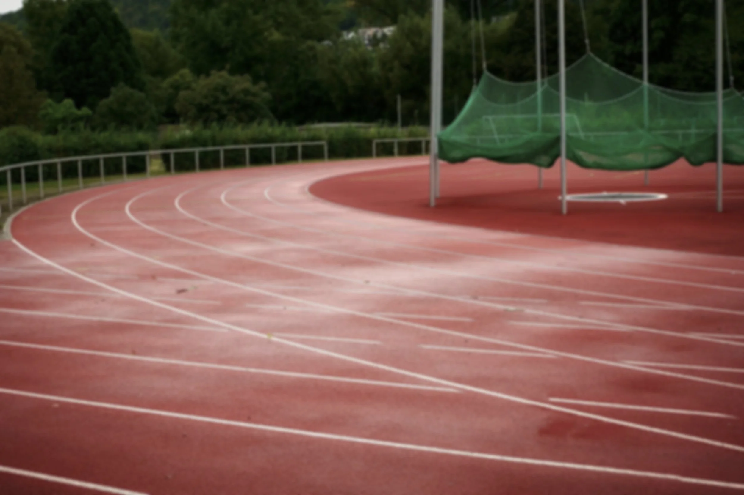 Empty outdoor running track with a green netted circle structure and a surrounding railing, set against a backdrop of trees and hills.