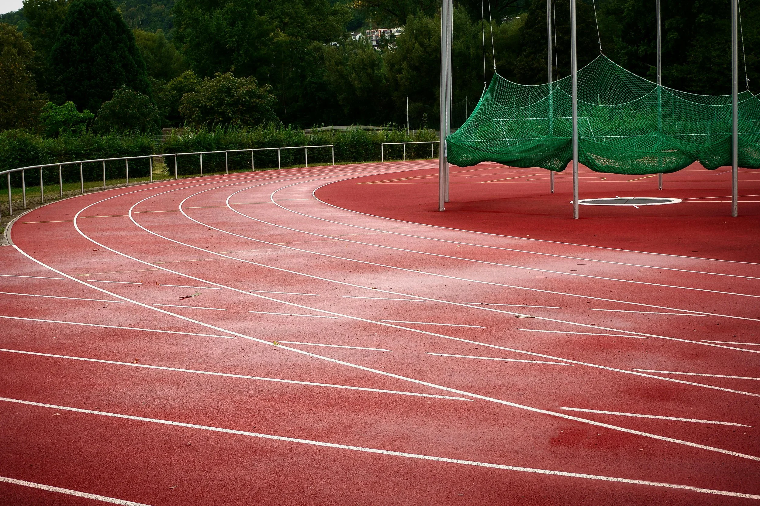 An outdoor running track with a green netted disc throw or hammer throw cage on the right side of the track, surrounded by lush green trees and hills in the background.