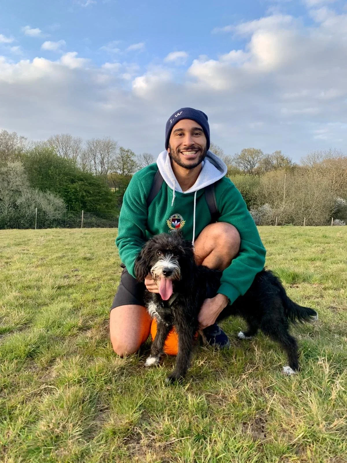 A young man kneeling on grass with a black and white puppy, smiling and outdoor landscape in the background.