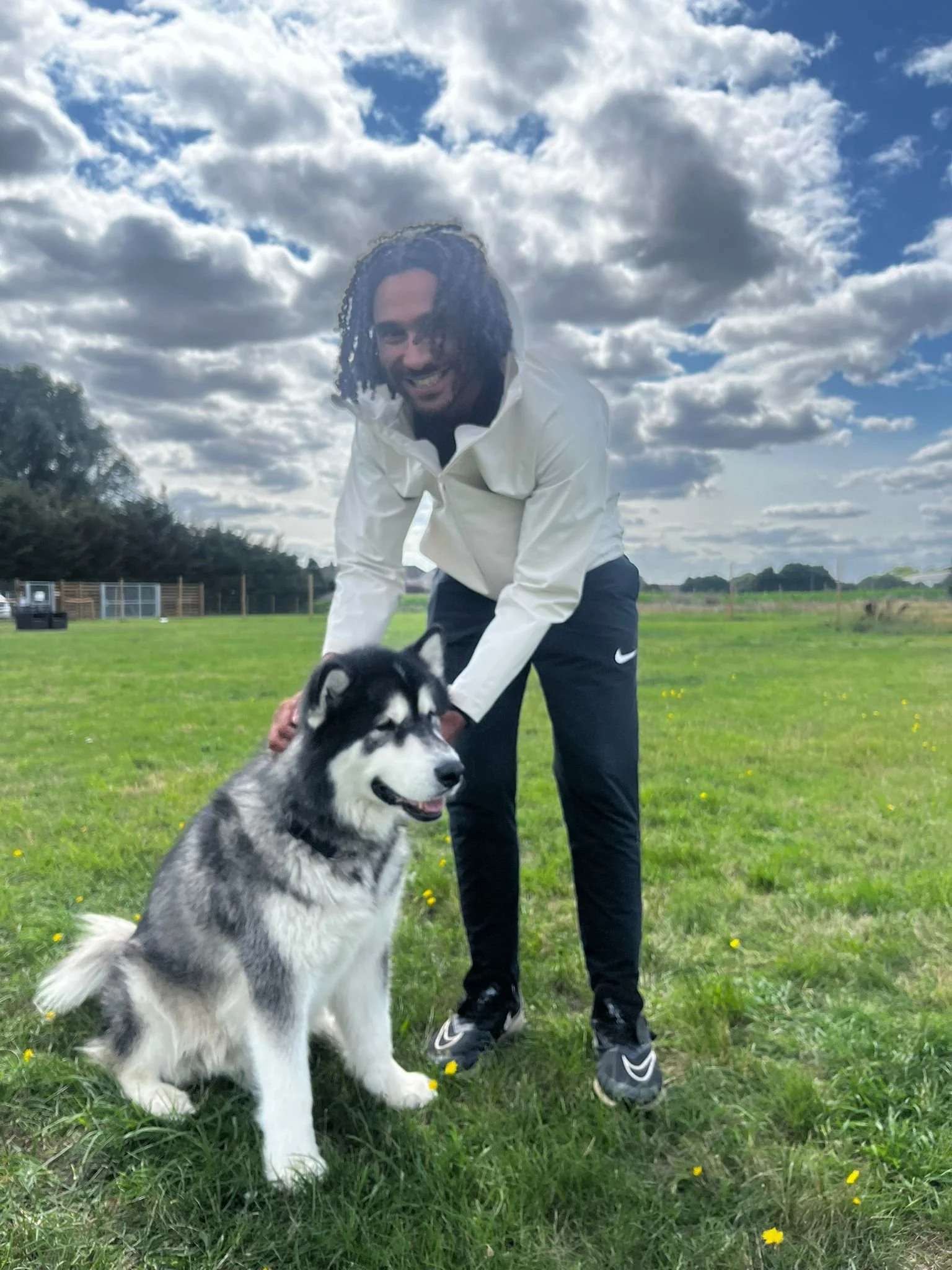 A smiling man with dreadlocks in a white jacket and black pants is outside in a grassy field with a Siberian Husky dog; the sky is partly cloudy.