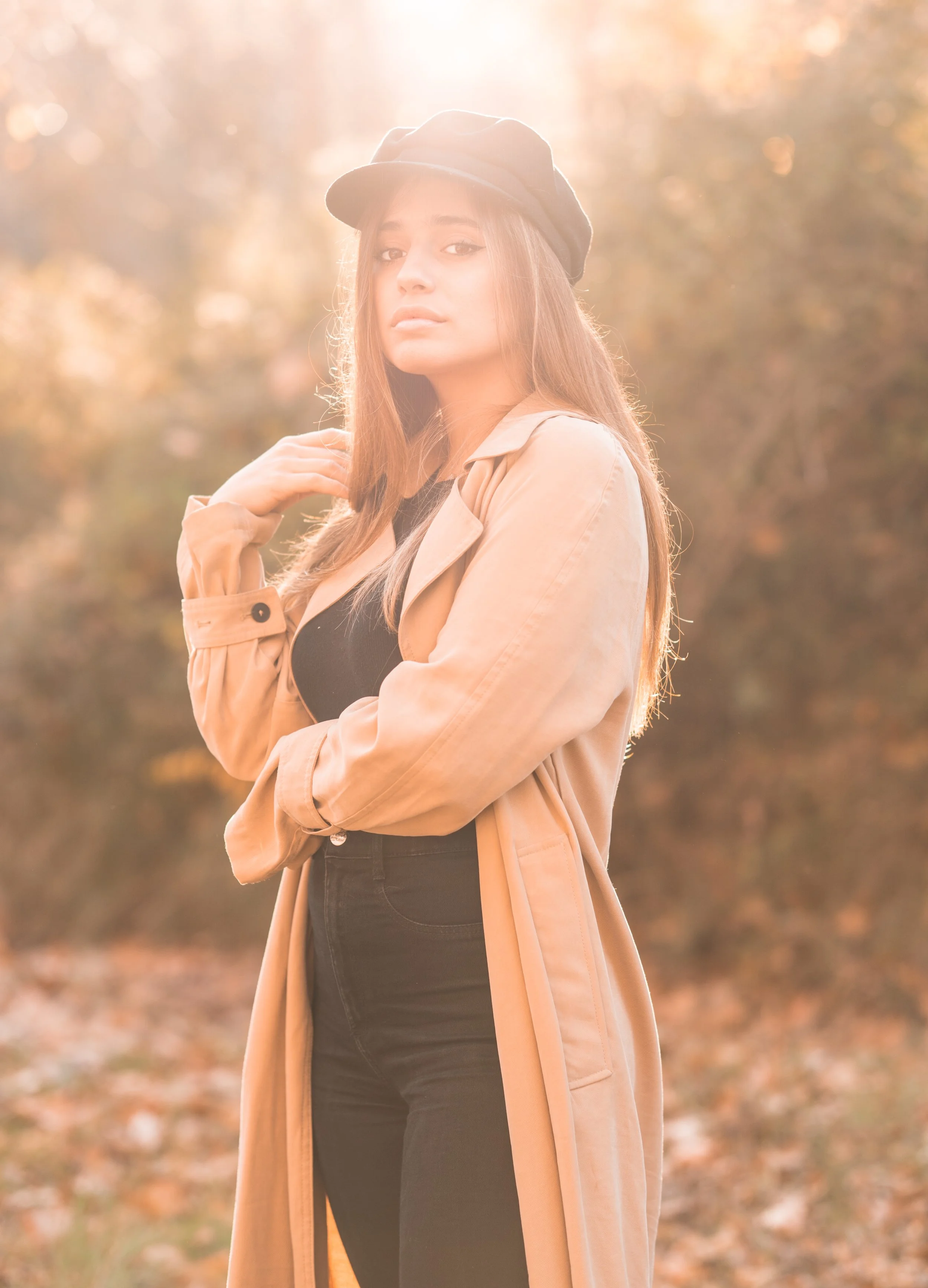 Joven mujer en un parque con árboles en otoño, vestida con abrigo beige, sombrero y ropa negra, posando con expresión pensativa.
