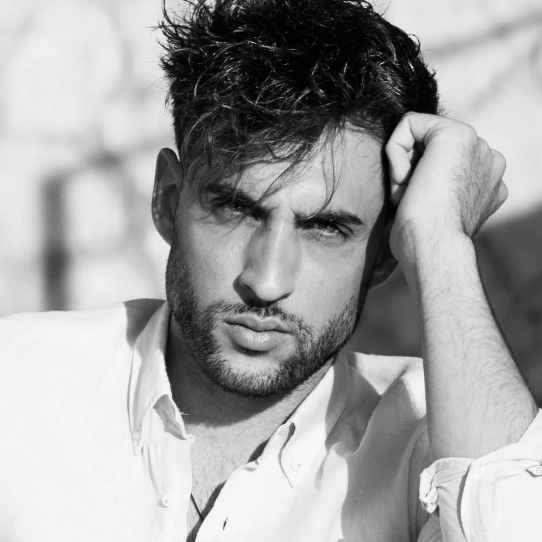 Retrato en blanco y negro de un hombre con cabello oscuro y barba, con expresión pensativa, descansando su cabeza sobre su mano, vestimenta casual con camisa blanca.