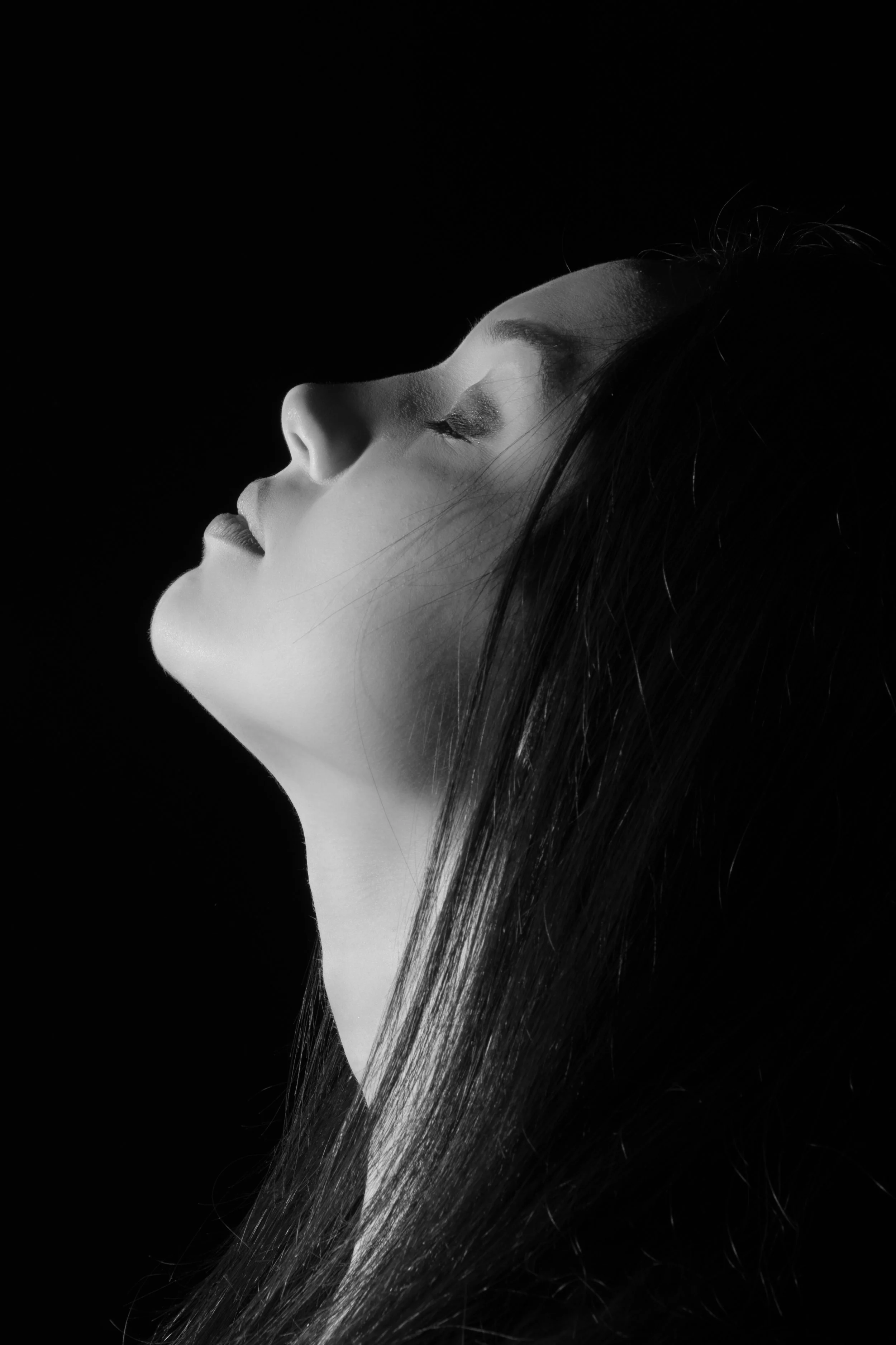 Retrato en blanco y negro de perfil de una mujer con los ojos cerrados y cabello largo y liso, con iluminación suave en un fondo negro.