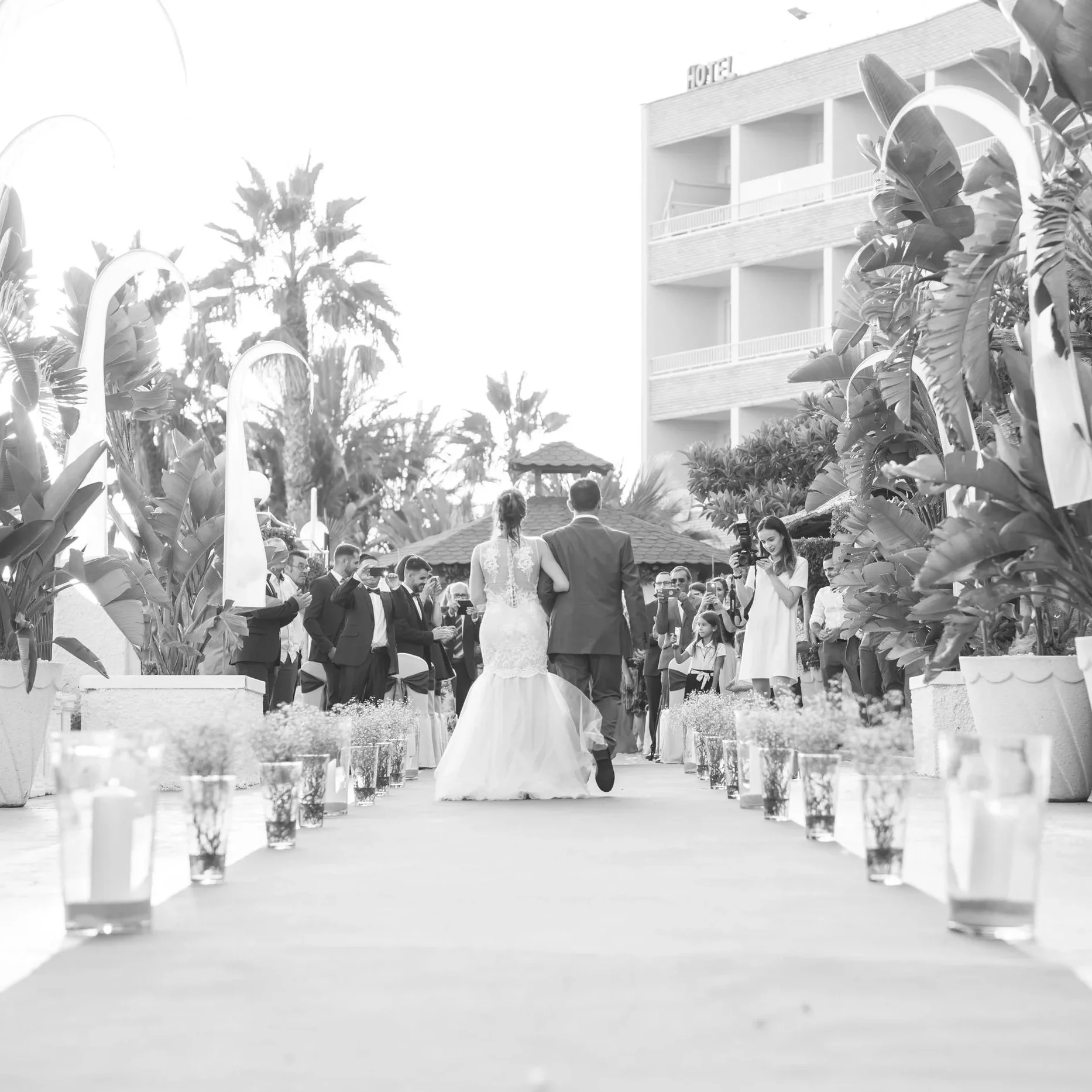 Boda al aire libre con pareja de novios caminando por un pasillo, rodeados de invitados, plantas tropicales y palmeras, en un ambiente con hotel en el fondo.