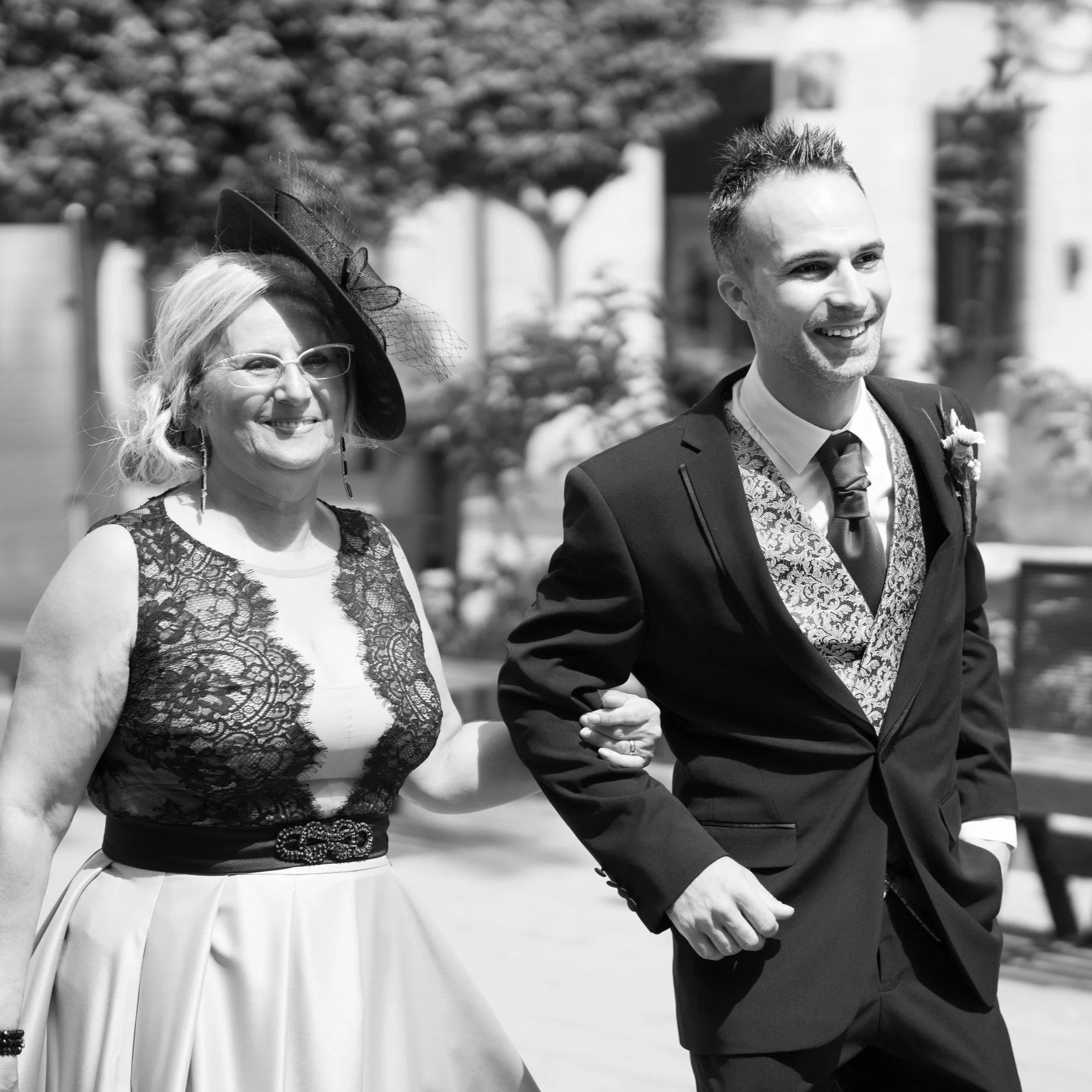 Una mujer y un hombre sonriendo mientras caminan juntos en un evento formal al aire libre, vestimenta elegante y ambiente urbano.