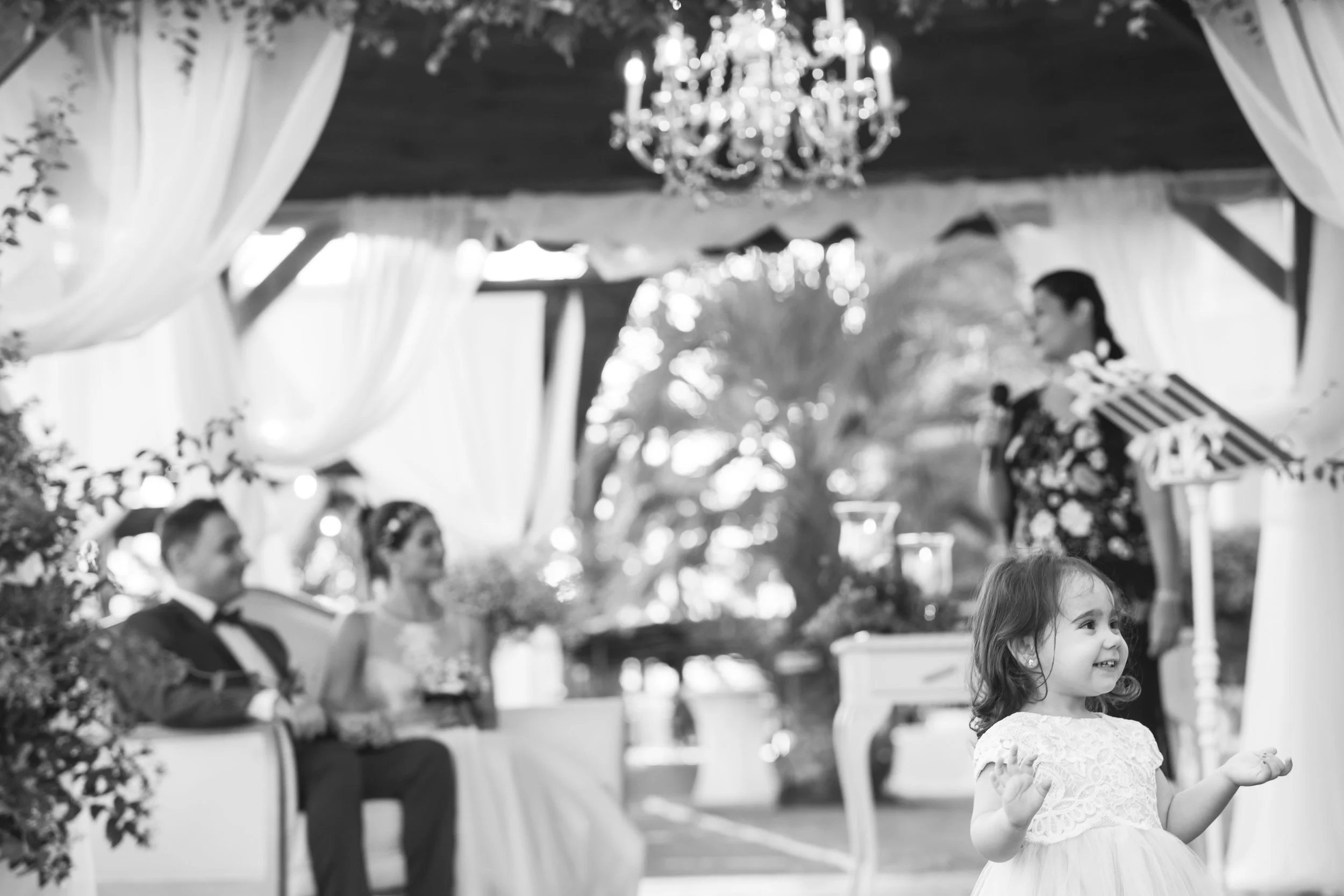 Niña sonriendo en un evento o celebración, con fondo de personas adultas sentadas en un escenario decorado con cortinas blancas y un candelabro.