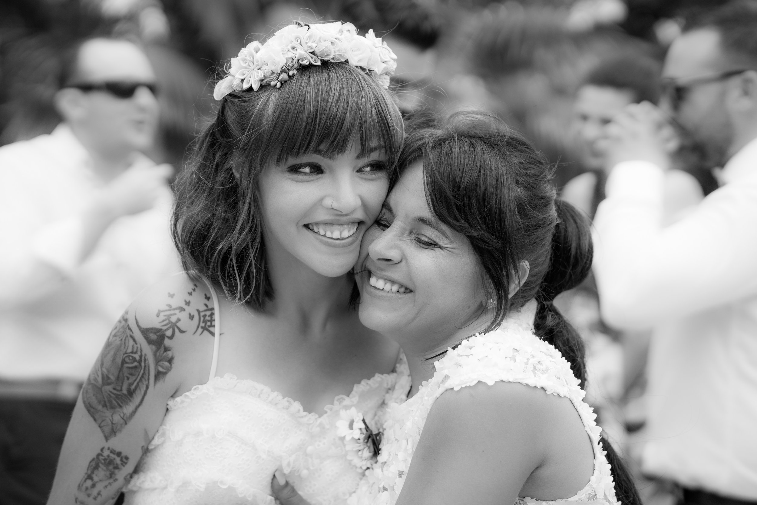 Dos mujeres abrazándose, una con una corona de flores y tatuajes en el brazo, sonriendo en una celebración al aire libre en blanco y negro.