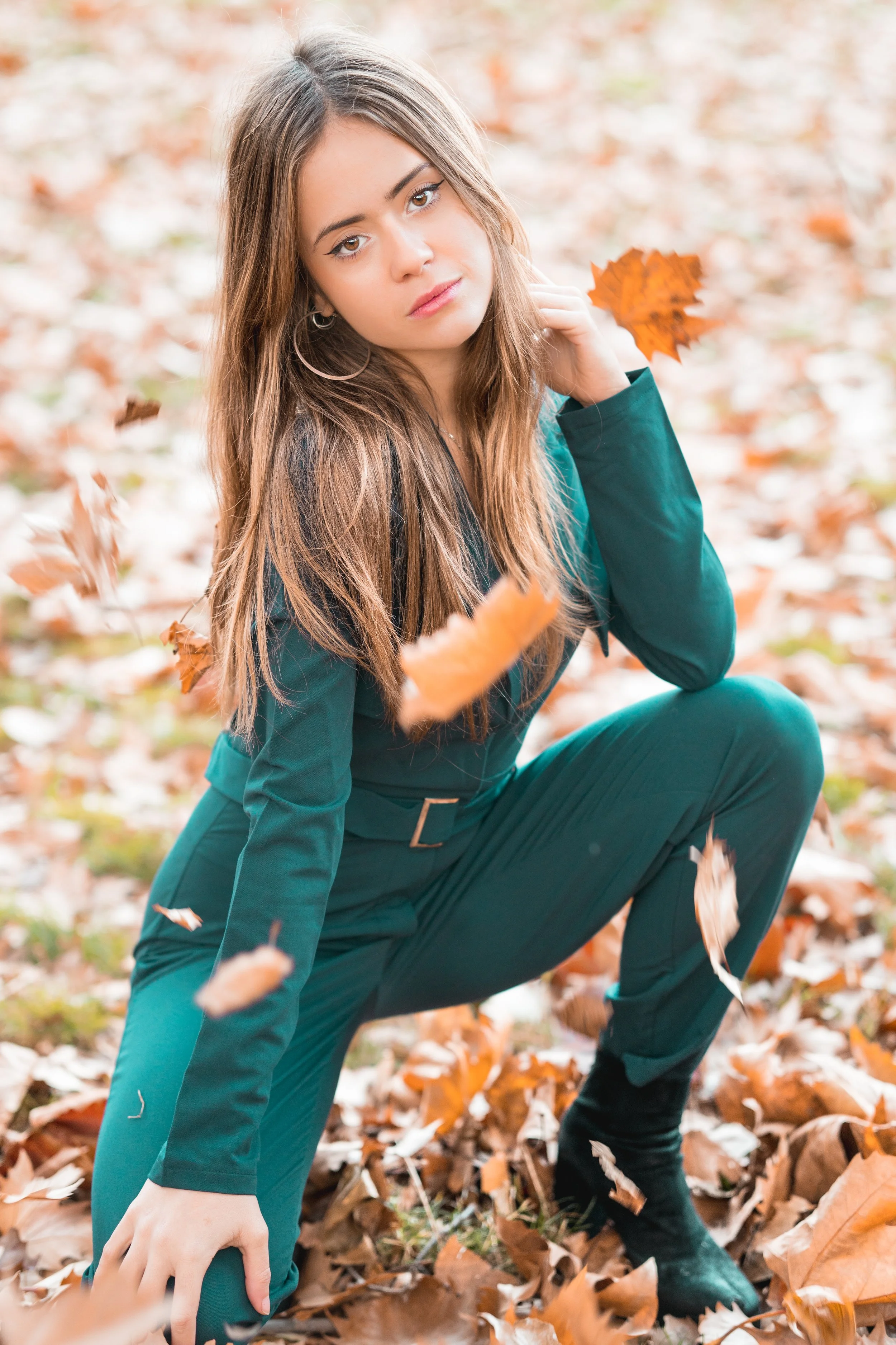 Una joven con cabello largo y ojos claros, vestida con un conjunto verde, está en cuclillas en un bosque cubierto de hojas otoñales, sosteniendo una hoja de color naranja en la mano y rodeada de hojas caídas.