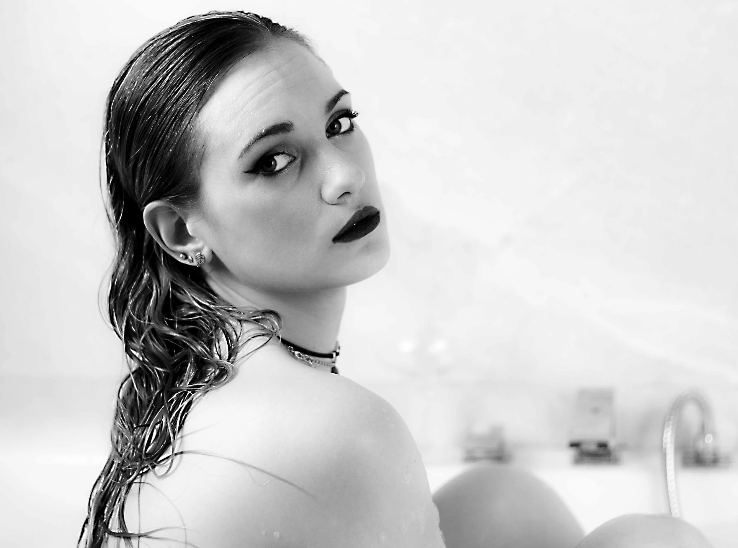 Mujer con cabello húmedo y maquillaje oscuro, mirando hacia la cámara, en un entorno probablemente de baño en blanco y negro.