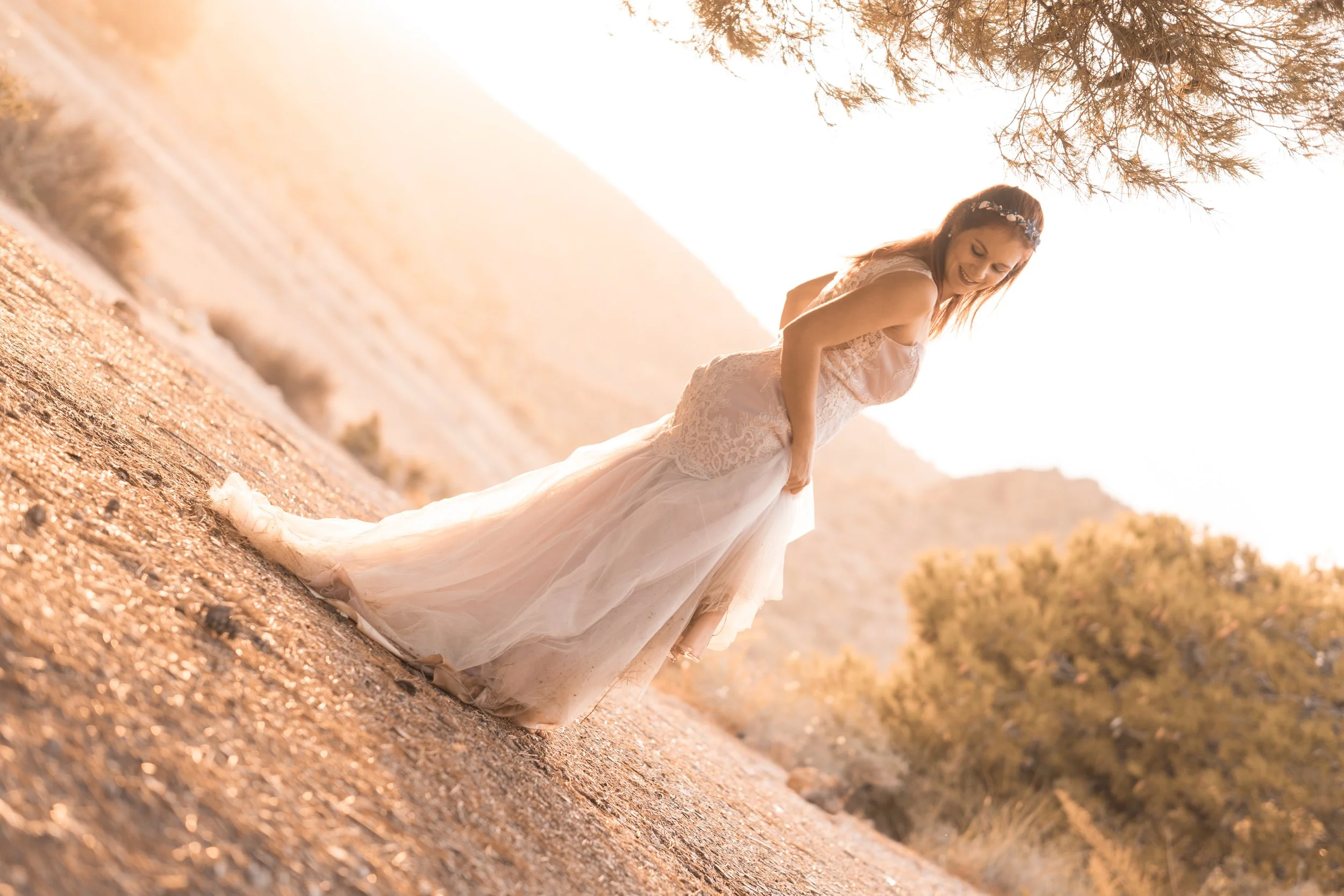 Mujer en vestido de novia en un paisaje árido al atardecer, sonriendo y mirando hacia abajo.