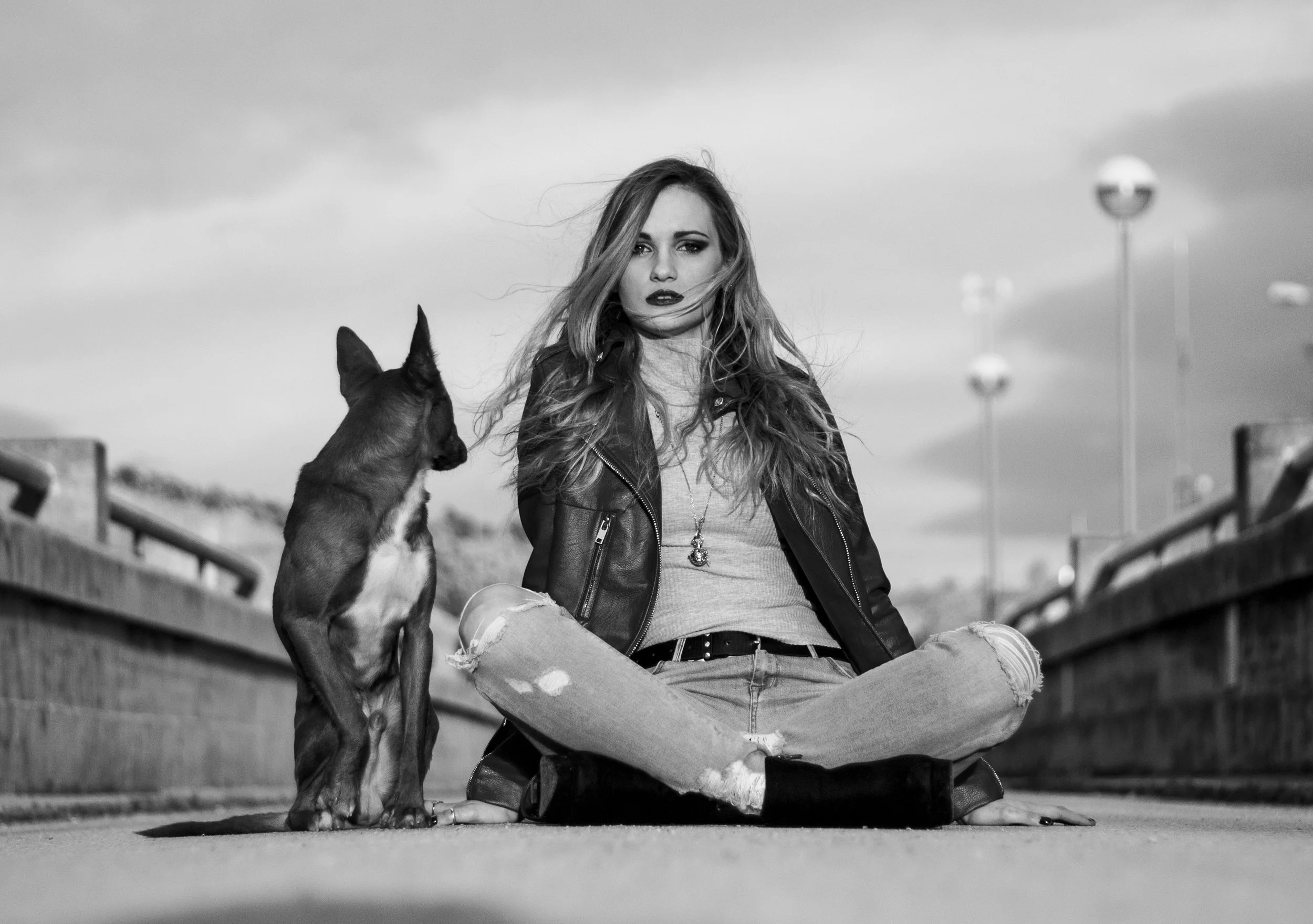 Joven mujer con cabello largo y suelto, vestida con chaqueta de cuero, jeans rotos y camiseta, sentada en el suelo de espaldas a una baranda en un puente, junto a un perro de raza pequeña, en un entorno urbano, todo en tonos negros y blancos.