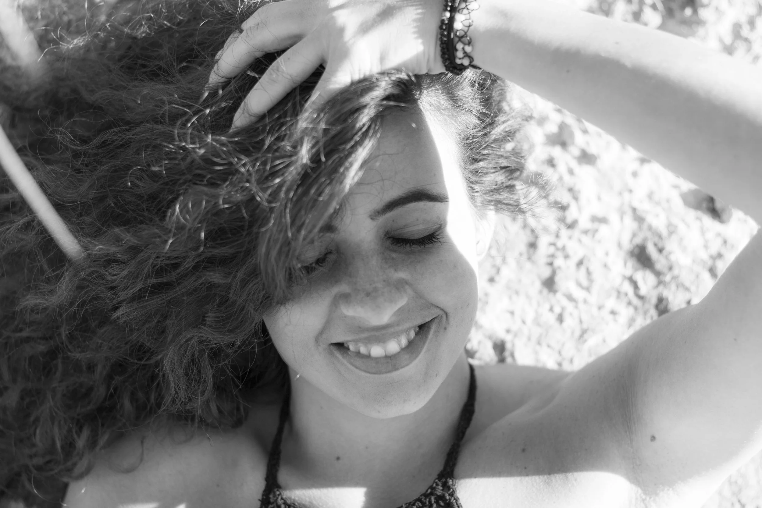 Mujer sonriendo con los ojos cerrados, acostada sobre una superficie arenosa, con cabello rizado y una mano en la cabeza.