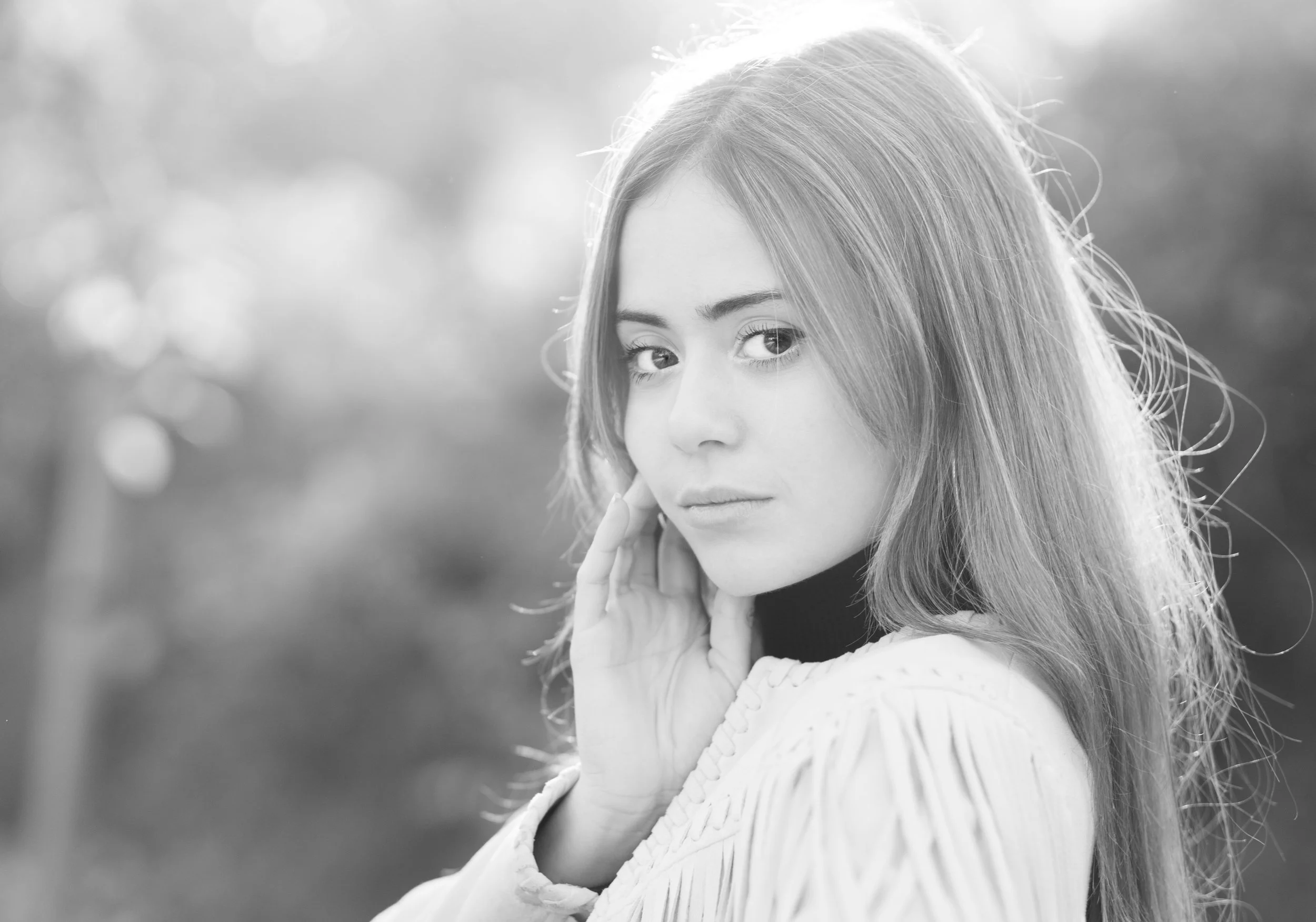 Retrato de una joven con cabello largo y liso, mirando hacia la cámara, tocándose la mejilla con una mano, fondo difuso en tonos claros, en blanco y negro.