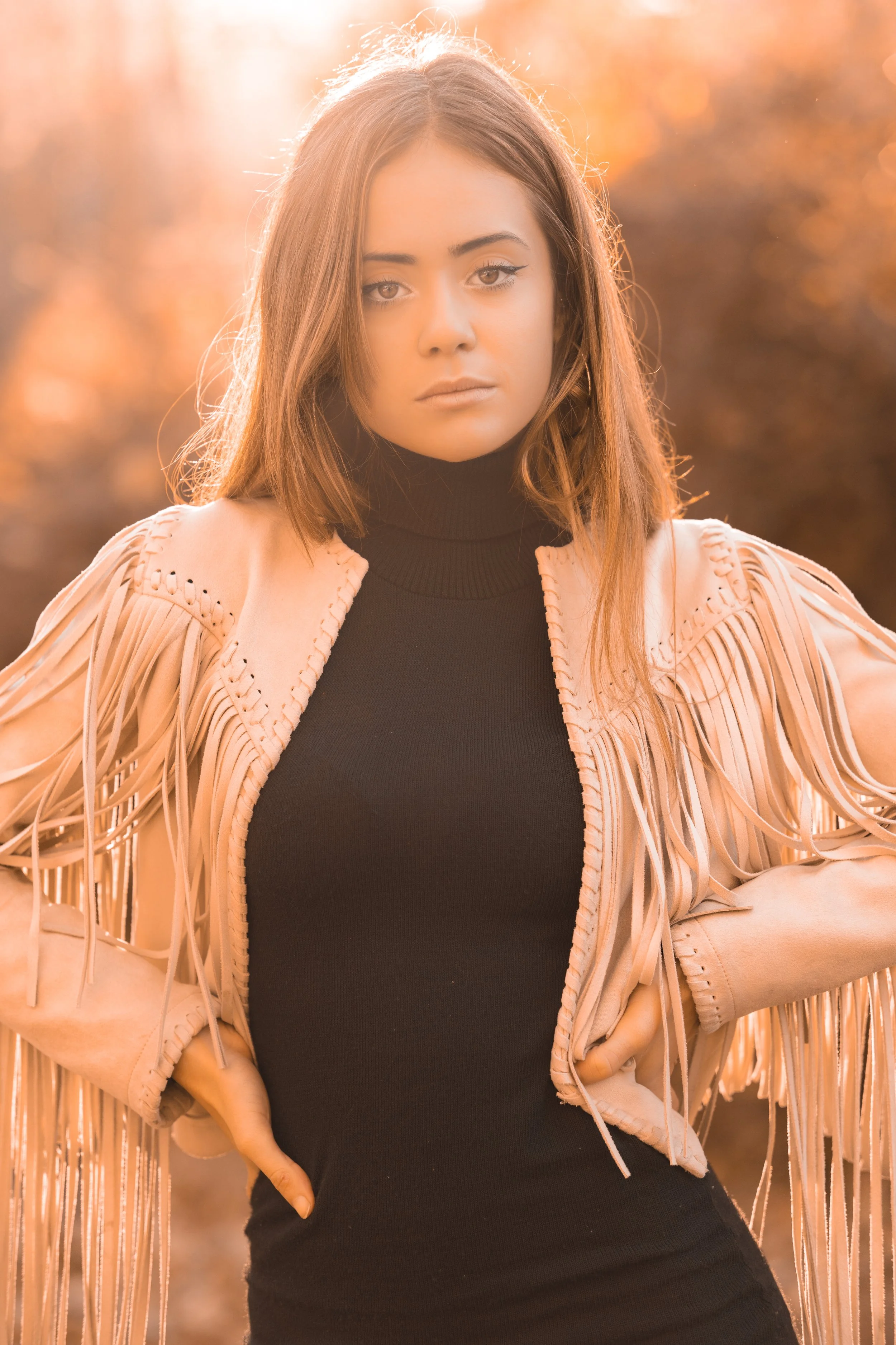 Joven mujer con cabello castaño, viste un suéter negro y una chaqueta con flecos, posando al aire libre con fondo difuso de tonos otoñales y luz solar