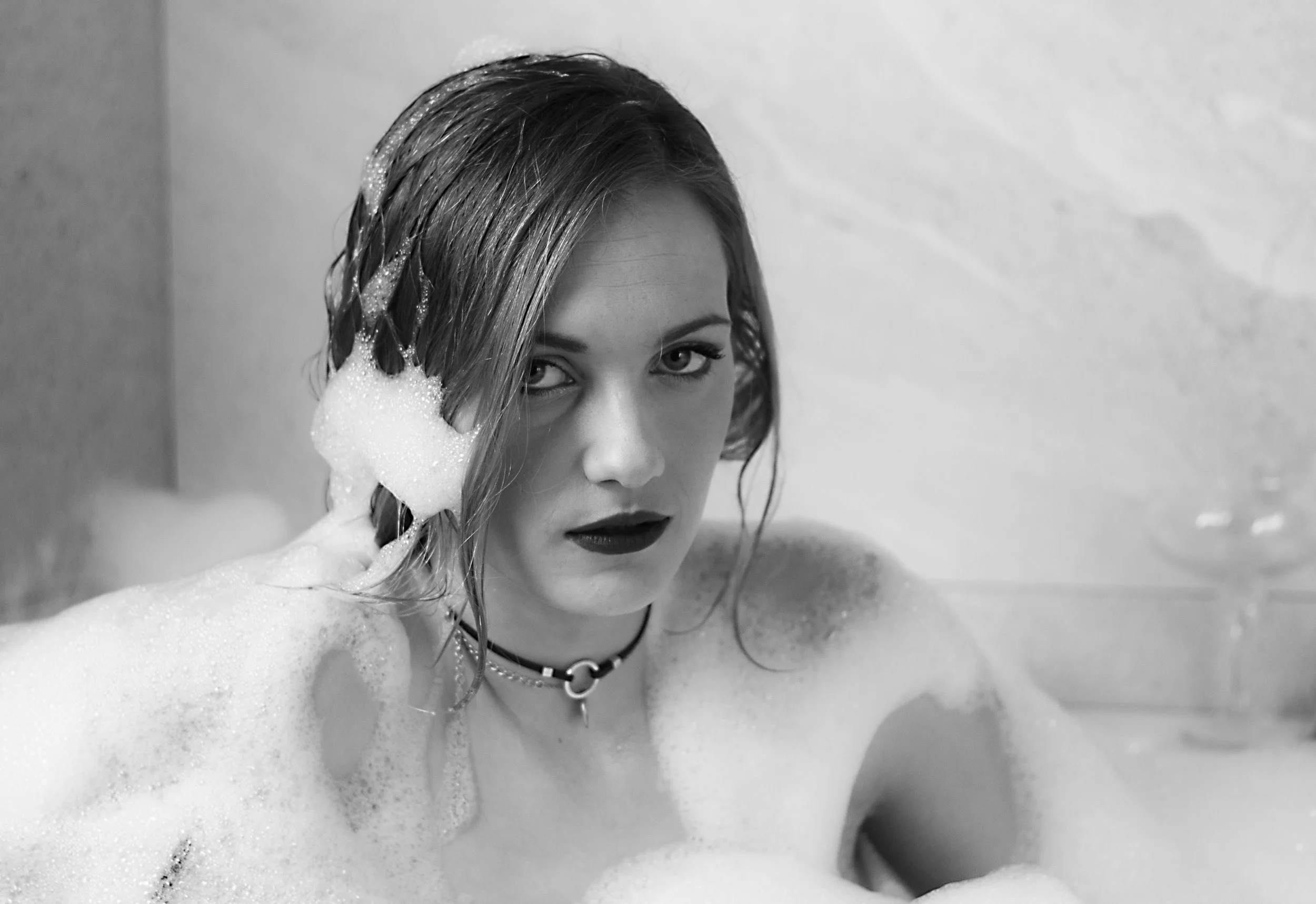 Mujer con cabello mojado y maquillaje oscuro, en una bañera con espuma, mirando hacia la cámara en un entorno de baño.