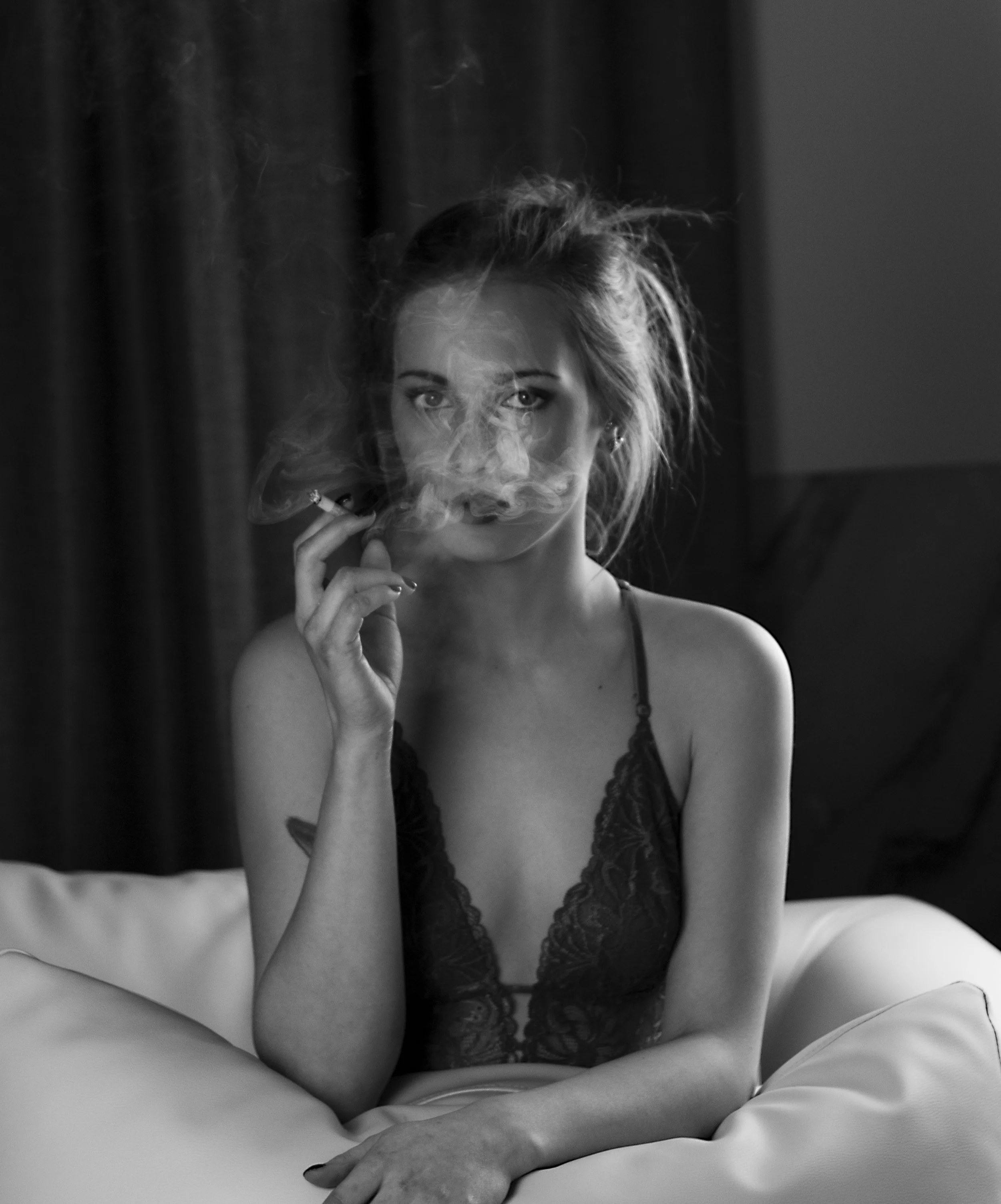 Una mujer joven de cabello despeinado y ojos intensos, vestida con lencería de encaje, está sentada en una cama y fuma un cigarrillo, generando humo en el aire, en una habitación con cortinas oscuras al fondo.