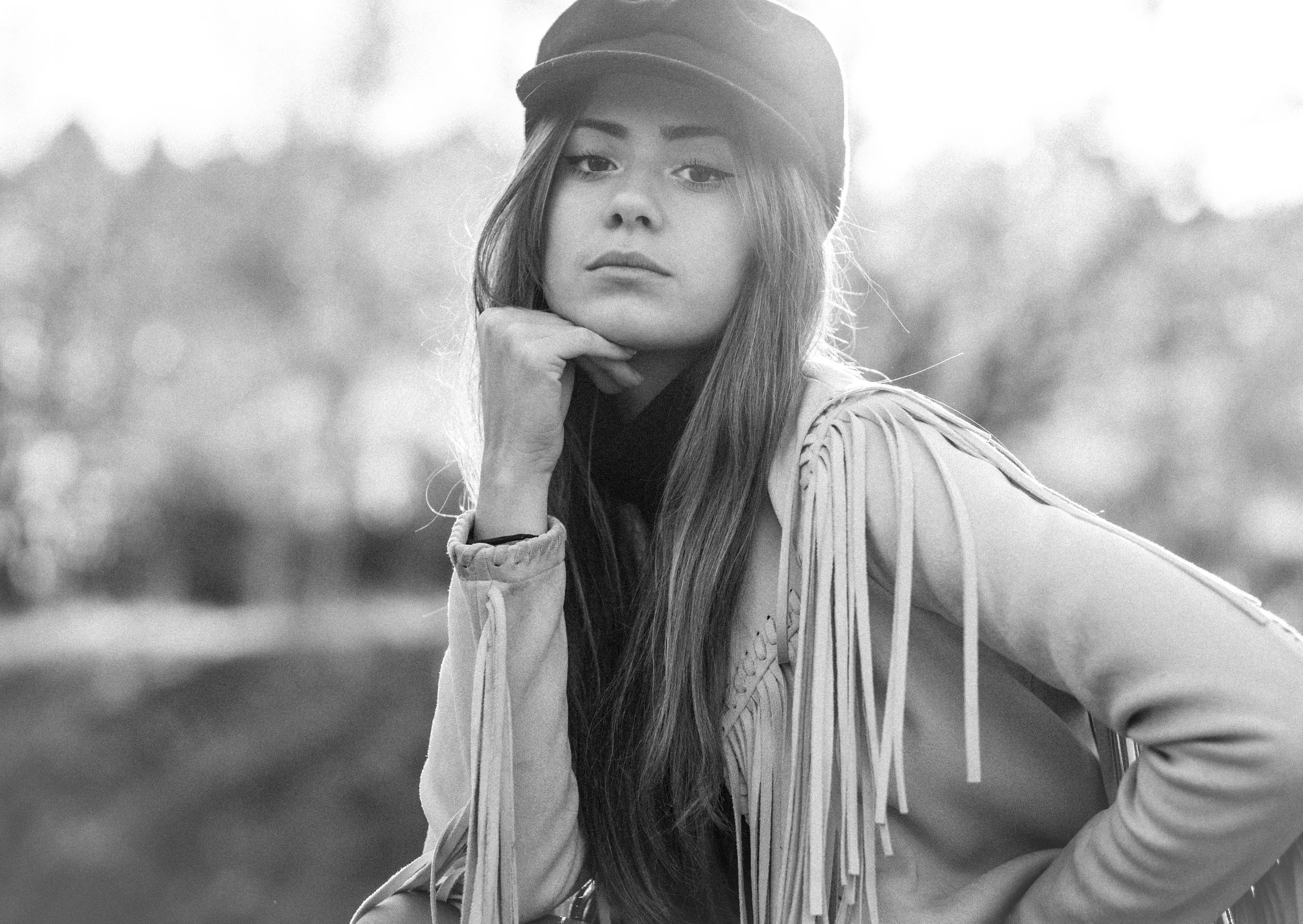Retrato en blanco y negro de una joven con sombrero, con cabello largo y una chaqueta con flecos, posando con la mano en la barbilla en un ambiente natural.