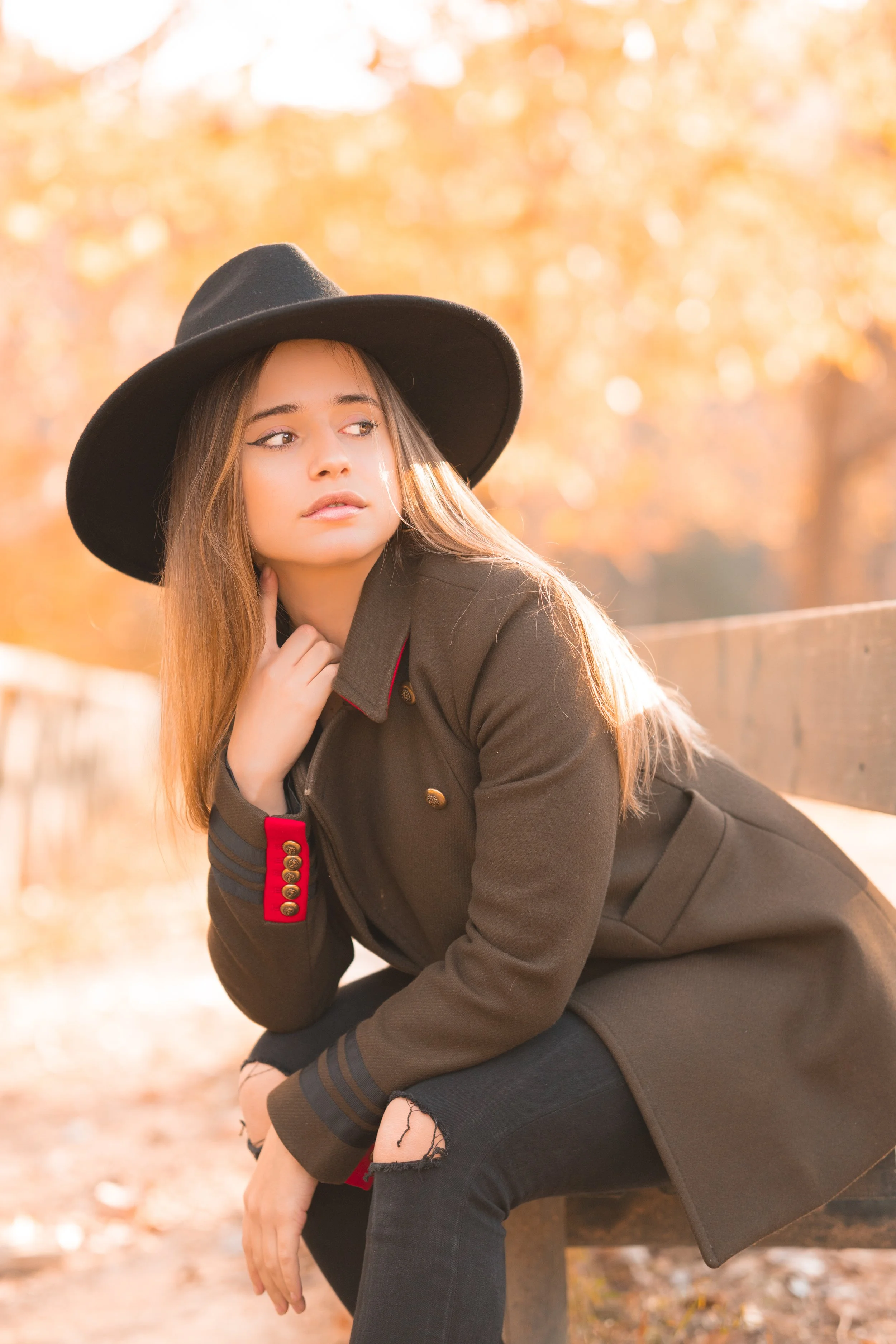 Joven mujer con sombrero negro y abrigo marrón, sentada en un banco en un parque en otoño.