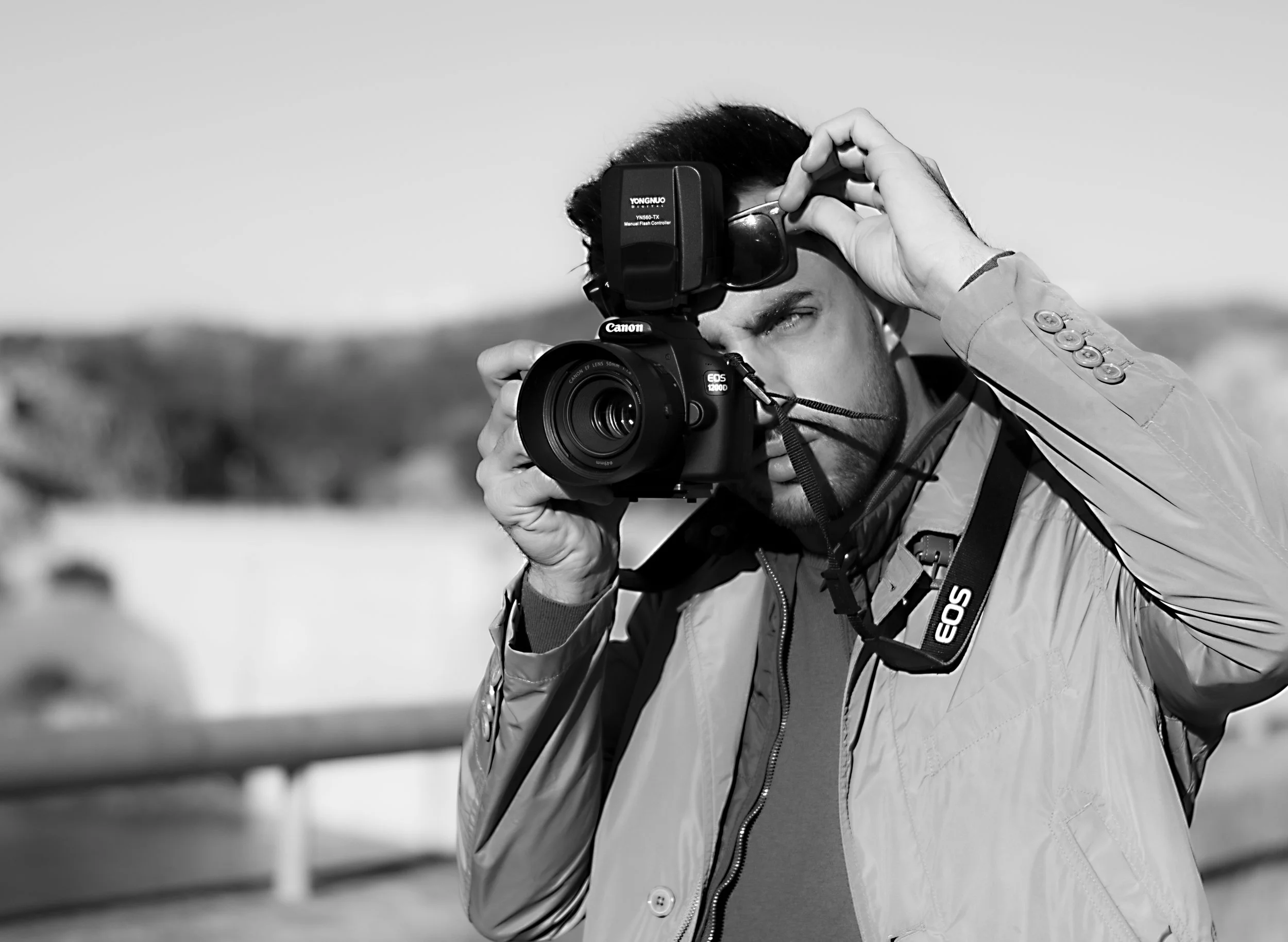 Hombre con cámara de fotografía en mano, ajustándose unas gafas, en un entorno al aire libre.