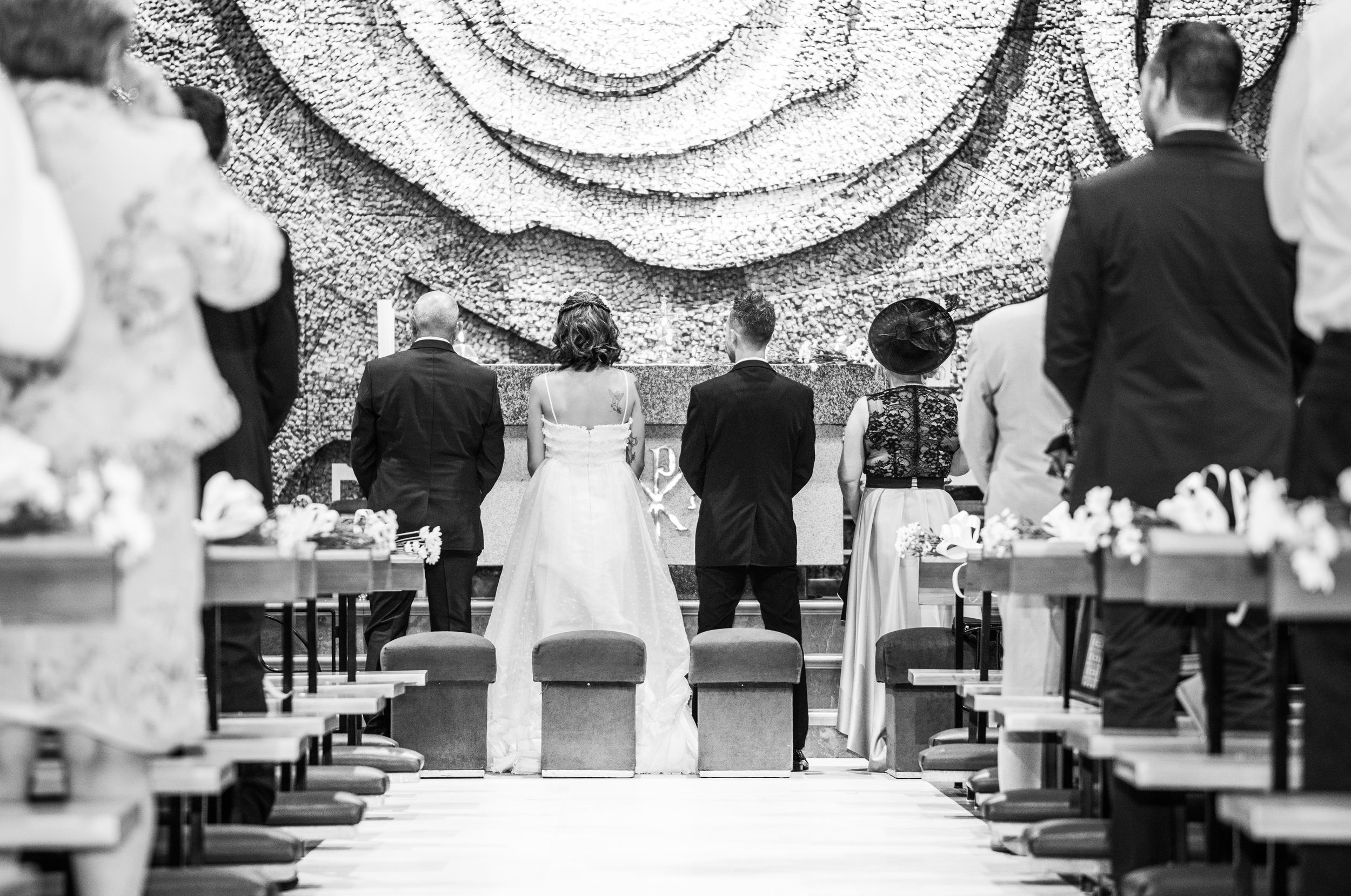 Una ceremonia de boda con varias personas, con una pareja en el centro, en un lugar con decoración floral y un fondo artístico detrás de los novios.