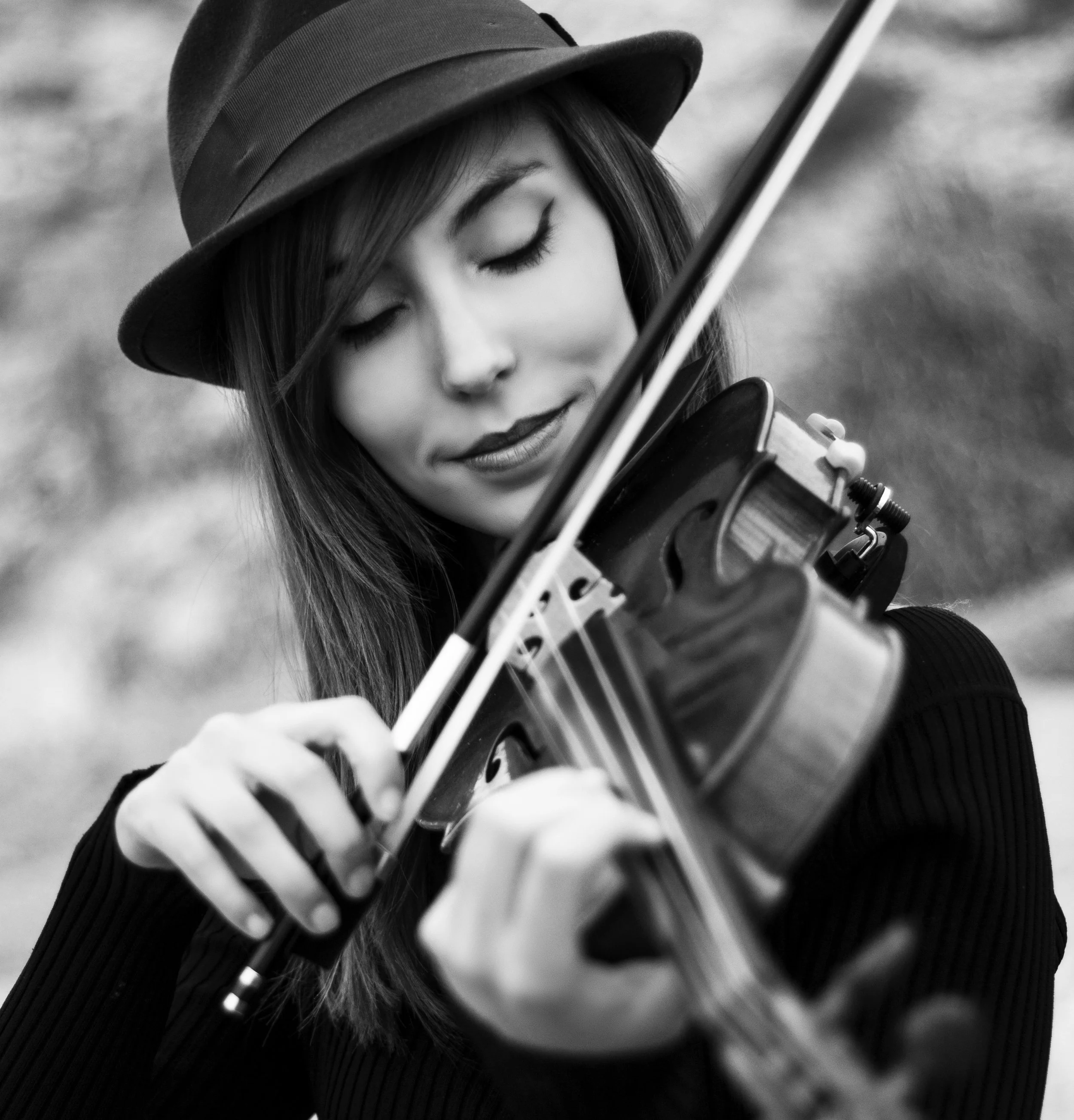 Mujer tocando violín, usa sombrero y ropa negra, con cabeza inclinada y ojos cerrados, en un fondo desenfocado.