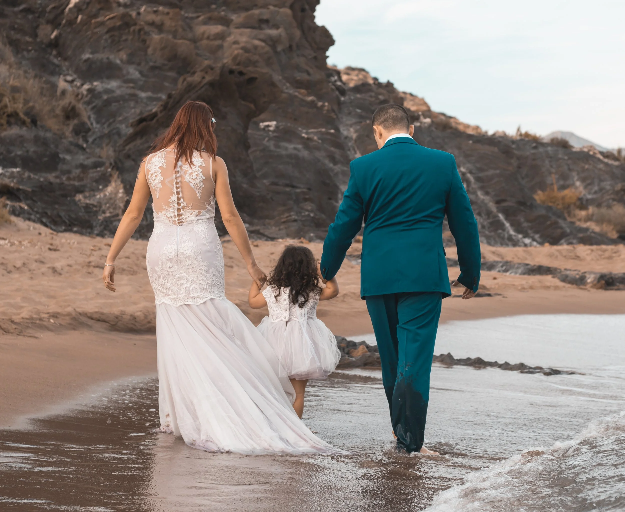 Familia con vestido de boda y traje caminando por la orilla del mar con una niña pequeña, en una playa con rocas y agua
