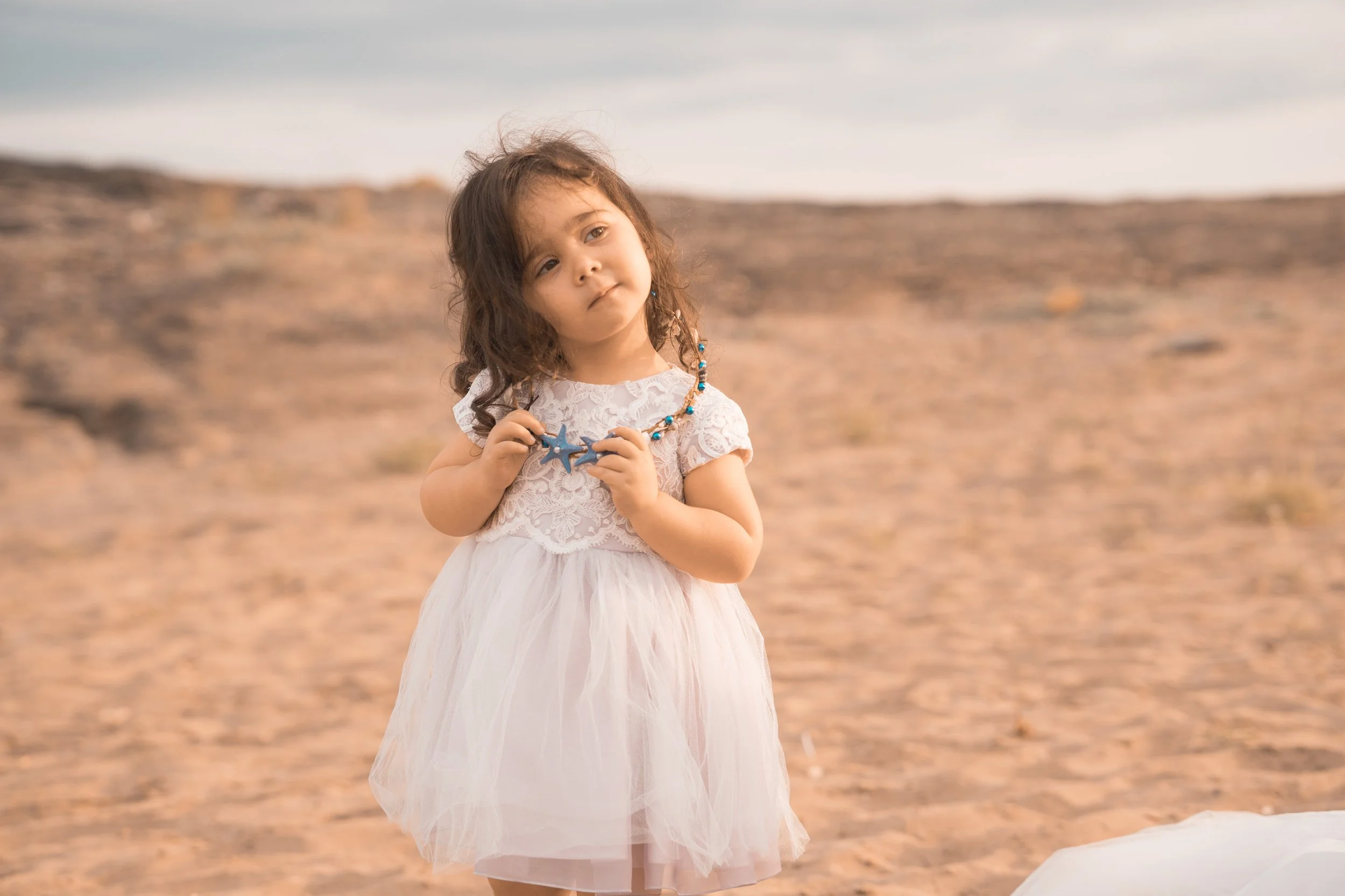 Niña pequeña con vestido blanco y collar de conchas, en un paisaje arenoso y desértico.