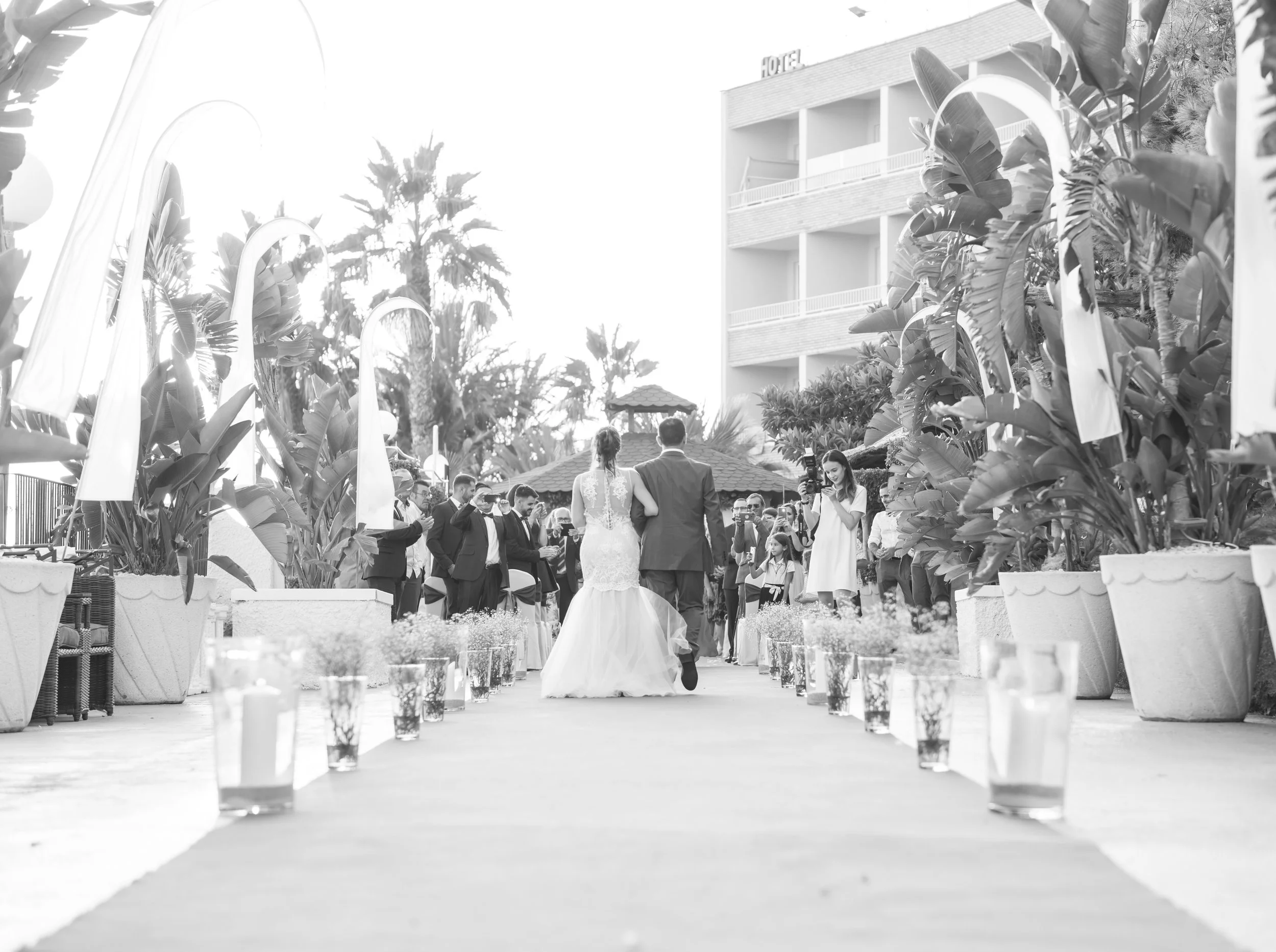 Boda en un espacio al aire libre con palmeras y decoraciones florales, con una pareja caminando por el pasillo y espectadores tomando fotos.