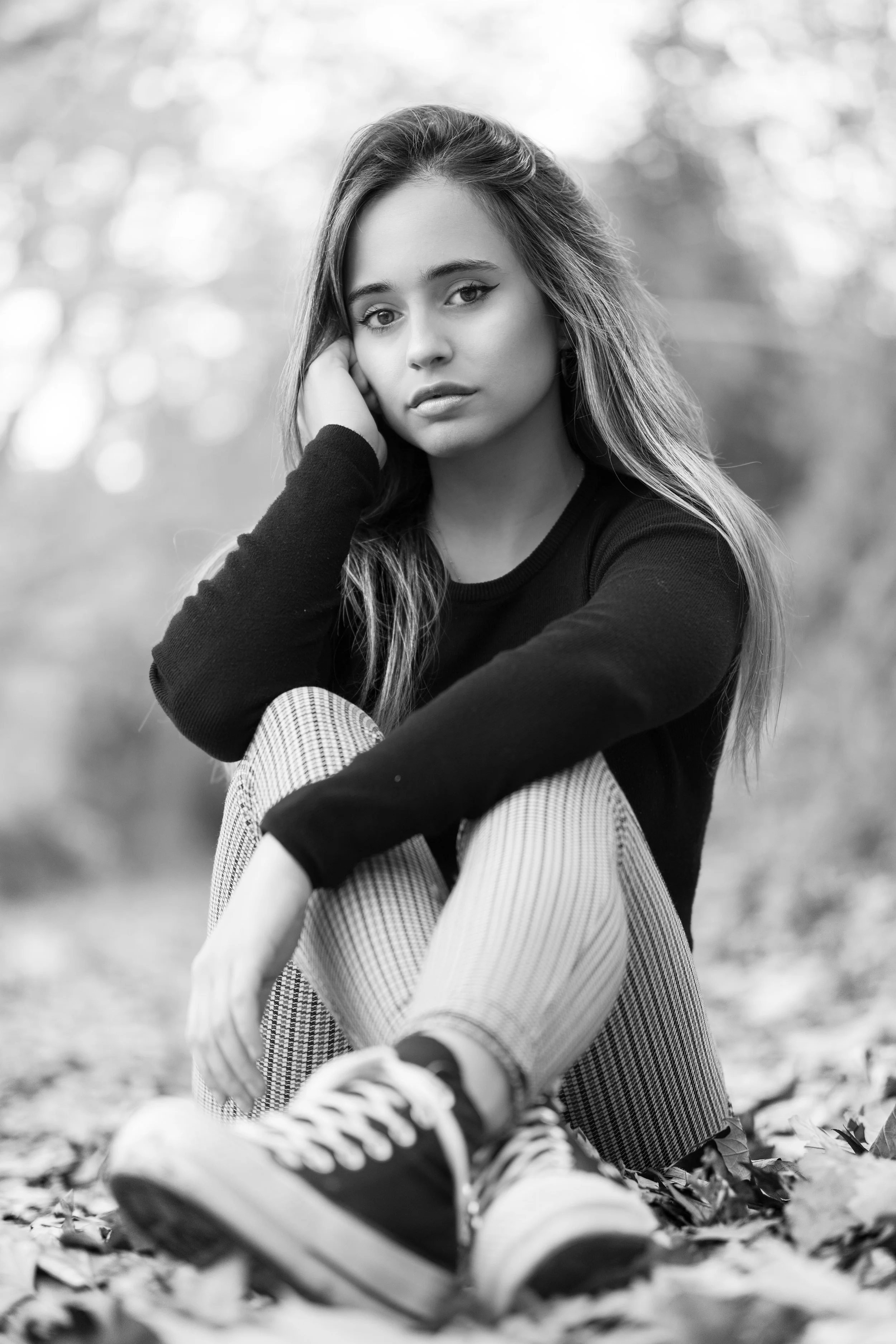 Retrato en blanco y negro de una joven mujer sentada en hojas, vestida con sudadera negra, pantalones de rayas y zapatillas deportivas, en un entorno natural al aire libre.