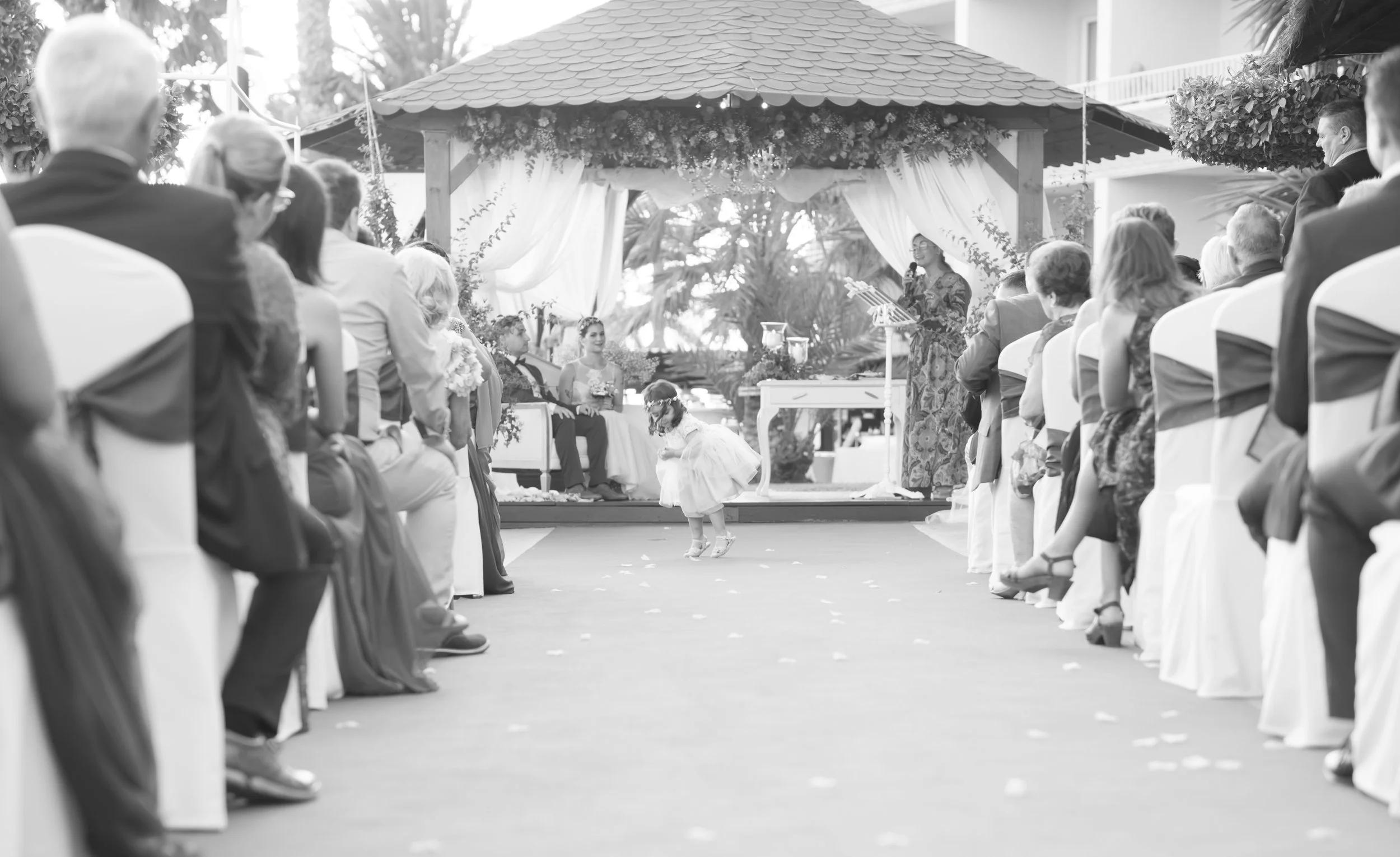 Una ceremonia de boda al aire libre con invitados sentados en ambos lados y una niña pequeña bailando en el centro, frente a un altar decorado con cortinas blancas y plantas.