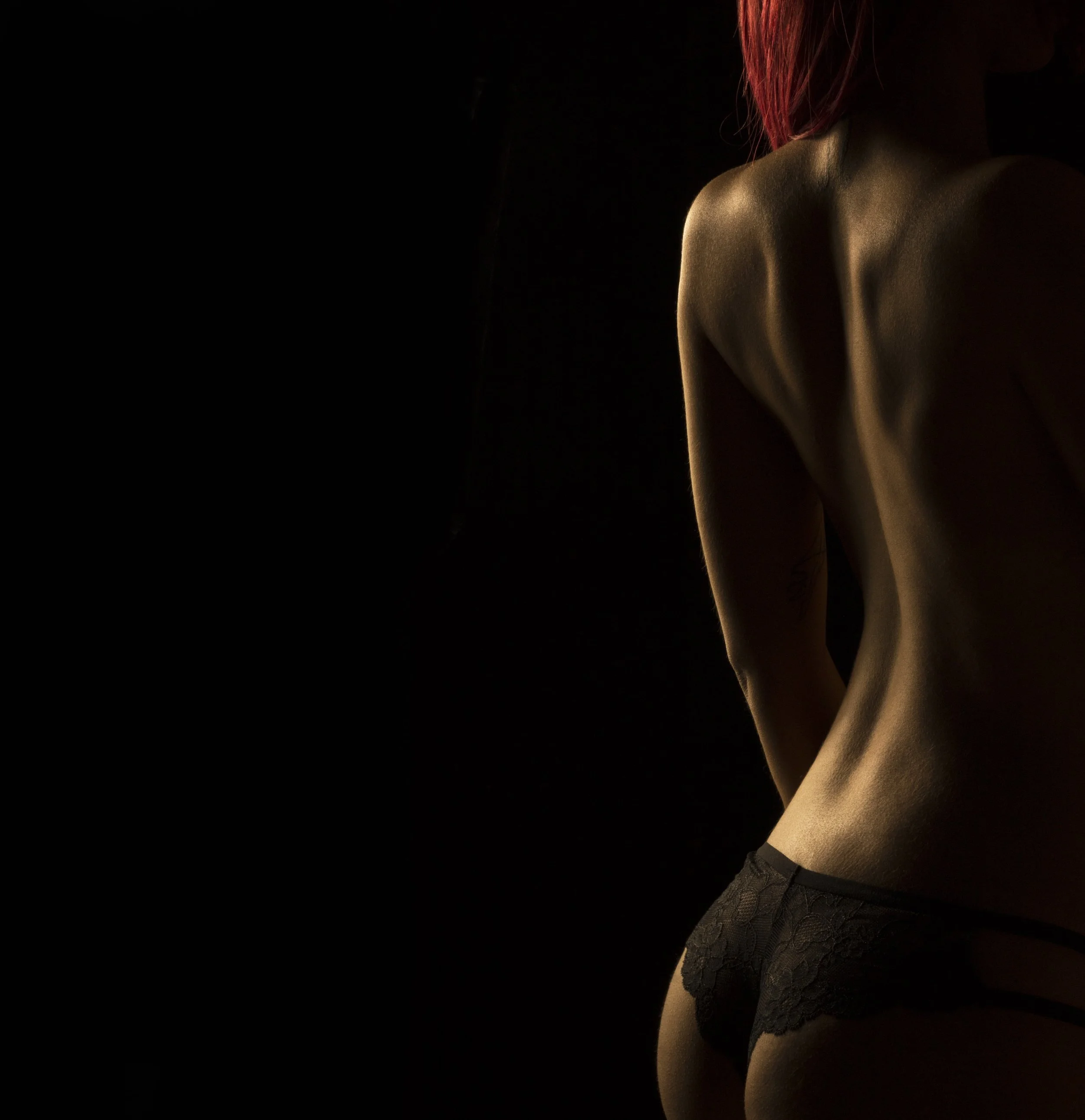 Mujer con cabello rojo y sin ropa, vista de espaldas, con ropa interior de encaje negro, en un fondo oscuro.