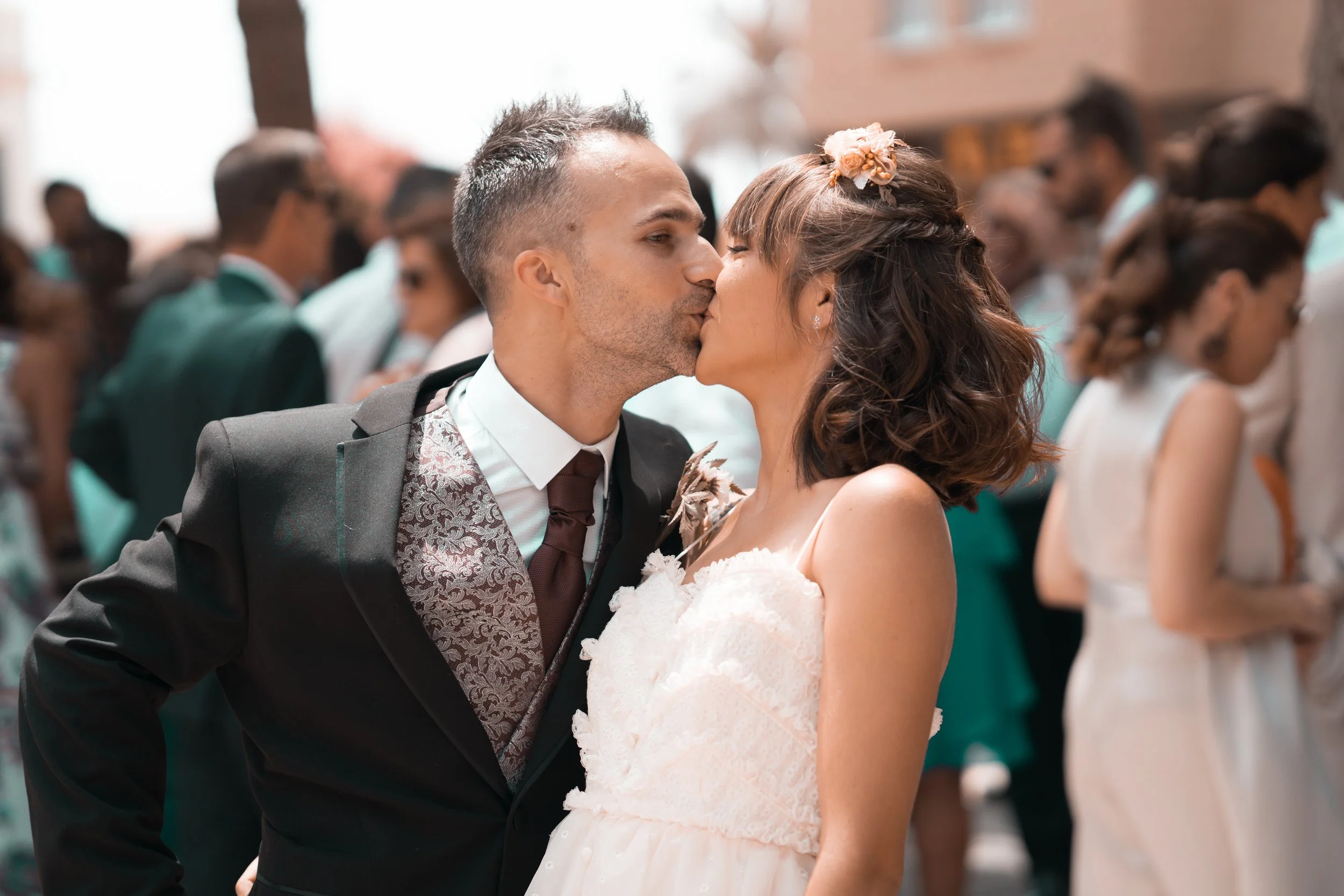 Pareja de novios besándose en una boda, rodeados de invitados en el fondo.
