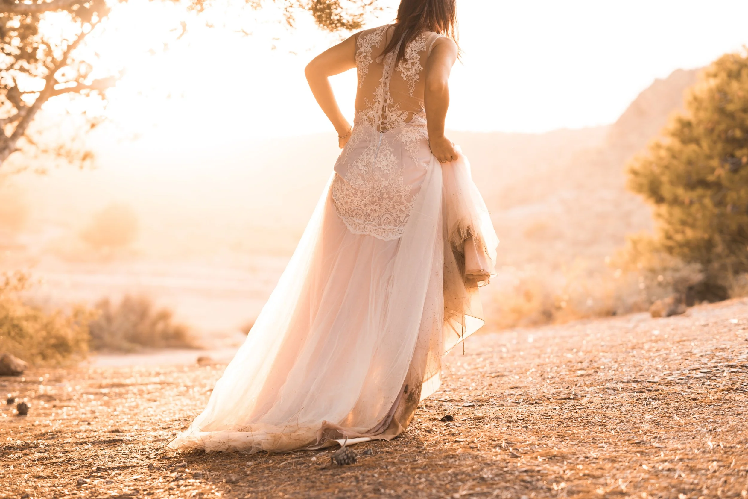 Mujer con vestido de novia, caminando en un sendero en un paisaje al atardecer, rodeada de árboles y montañas.