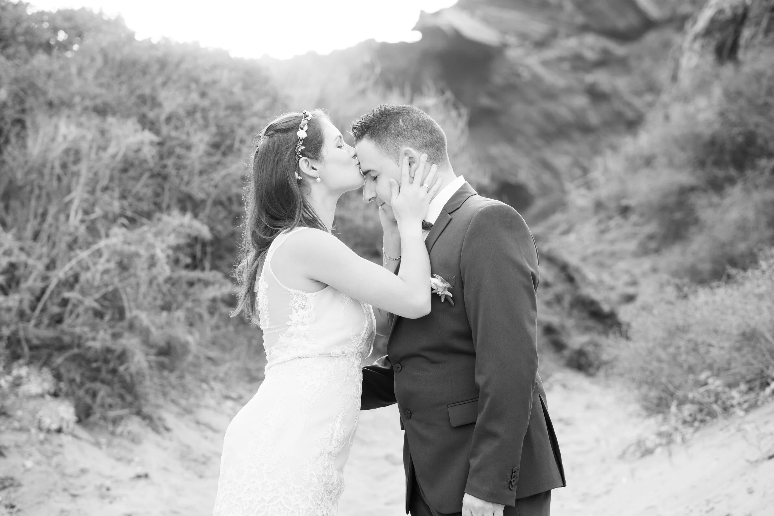 Una pareja de novios en un abrazo en la playa, la mujer le da un beso en la frente al hombre en una foto en blanco y negro.