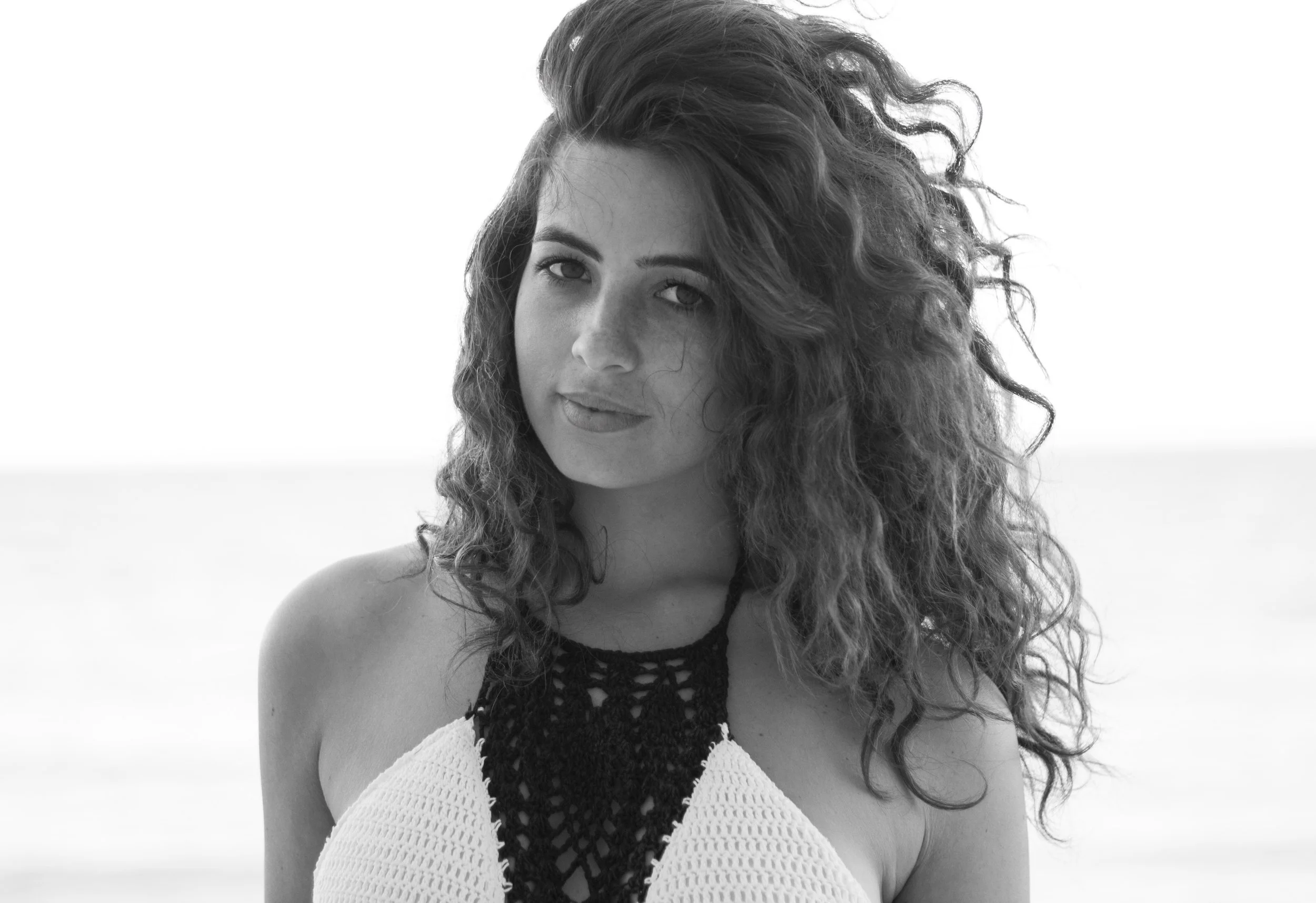 Retrato en blanco y negro de una mujer con cabello rizado y expresión serena, usando un top de crochet en la playa