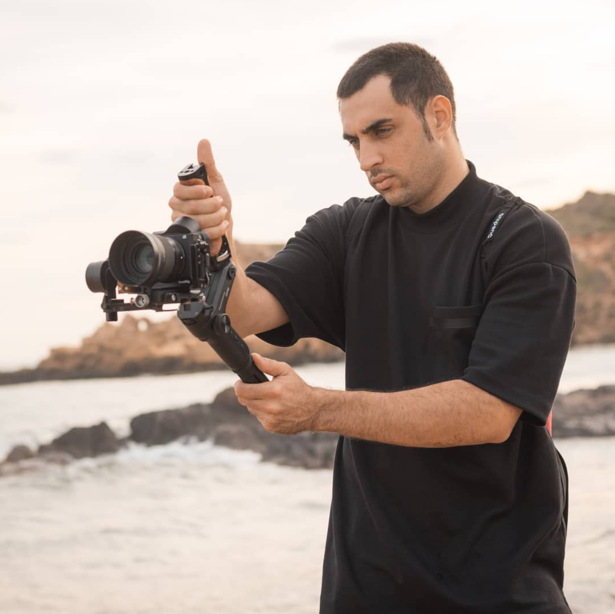 Hombre ajustando un estabilizador de cámara en una playa con rocas y agua