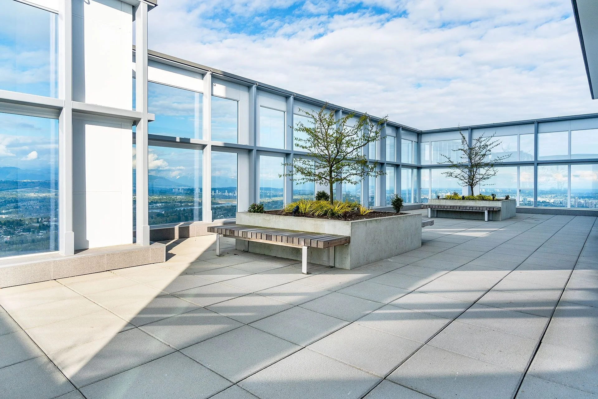 Rooftop terrace with large glass windows, potted trees, and city view, with partly cloudy sky