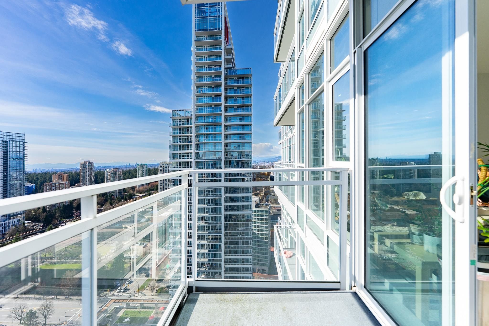 View of a city skyline with modern high-rise glass buildings from a balcony with glass walls and white railings, under a clear blue sky with some clouds.