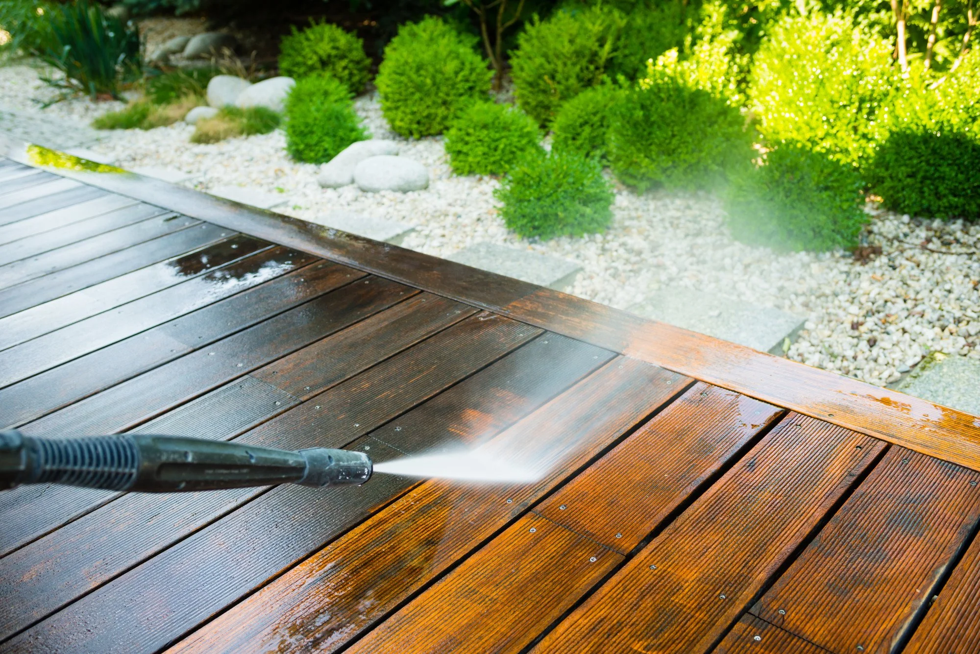 A power washer cleaning brown wooden deck with water spray, with green bushes and decorative white rocks in the background.