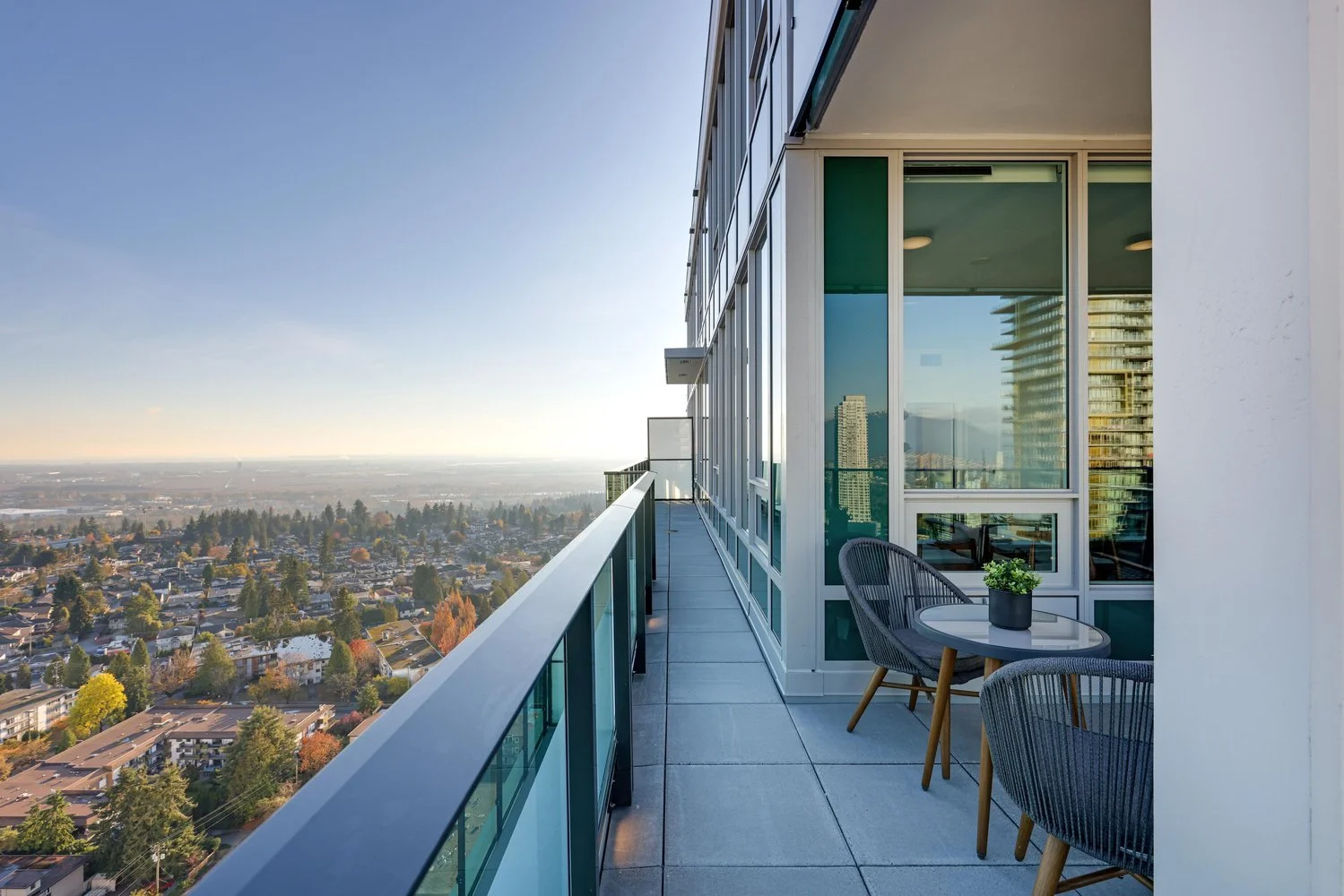 View from a high-rise balcony overlooking a cityscape with buildings, trees, and mountains in the distance during sunset. The balcony has a small table with two chairs and a potted plant.