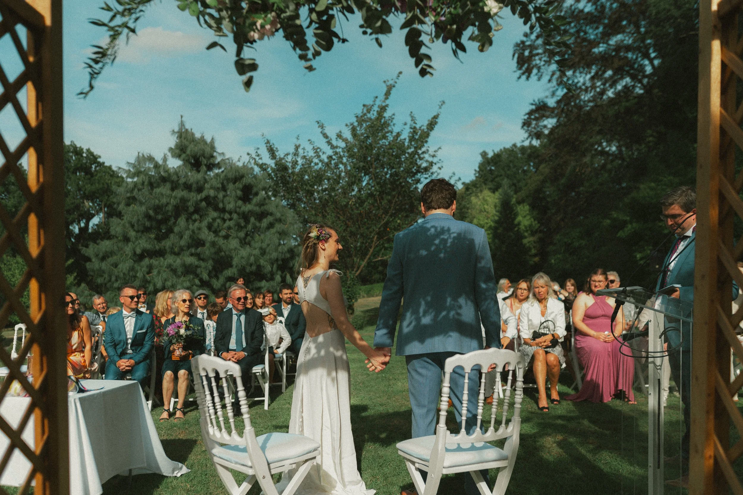 Un couple de mariés se tenant la main lors de leur mariage en plein air, entouré d'invités assis sous un ciel bleu avec des arbres en arrière-plan.