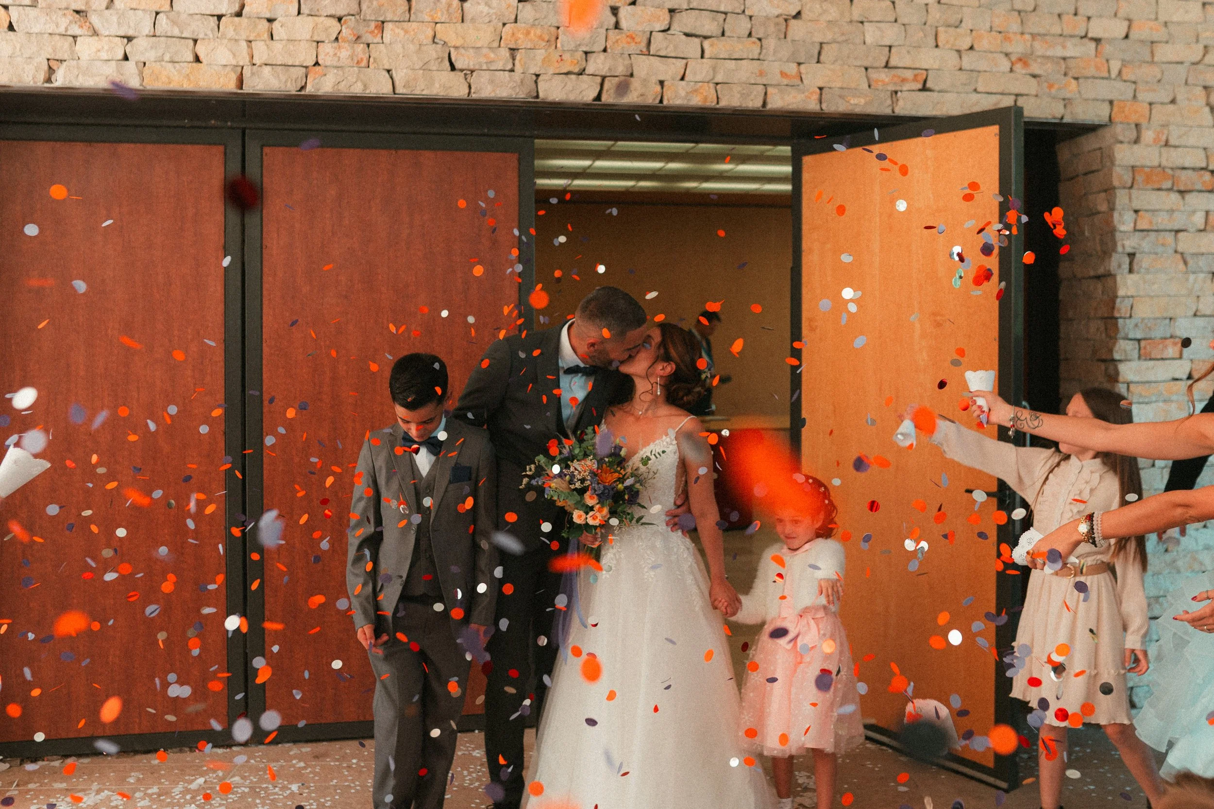 Un couple de mariés s'embrassant sous des confettis lors de leur réception, entourés de deux enfants et d'autres invités célébrant derrière une porte ouverte avec des murs en pierre