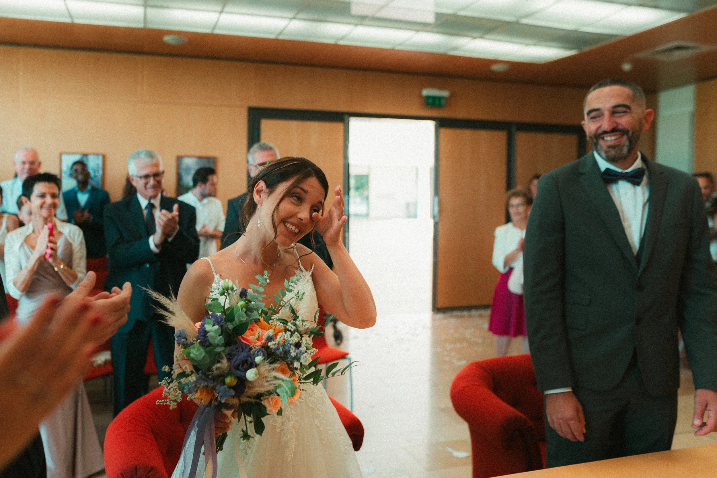 Un couple de mariés lors de leur mariage, entourés de leurs invités à l'intérieur d'une salle, la mariée en robe blanche tenant un bouquet de fleurs et le marié en costume, tous deux souriants.
