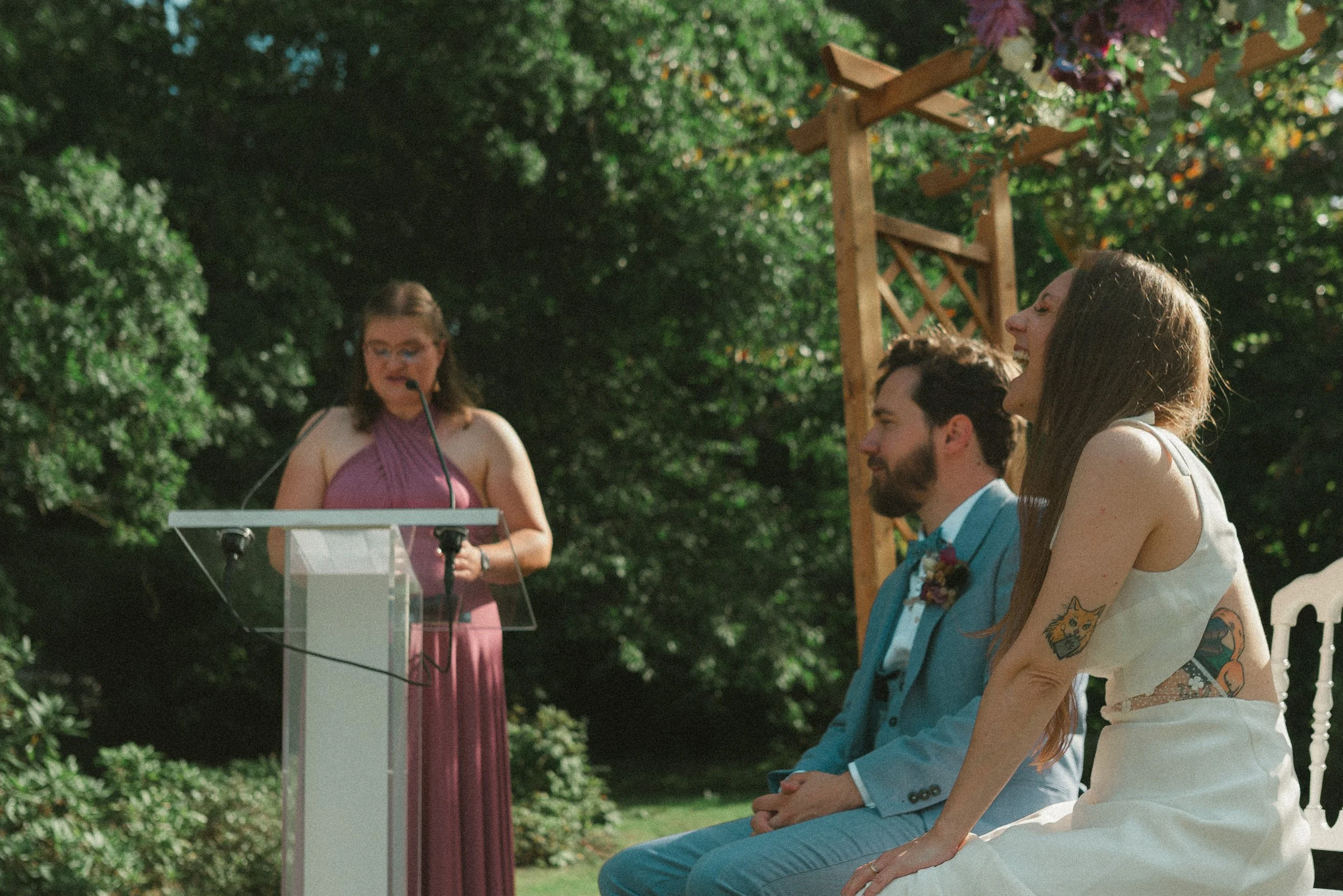 Une cérémonie en plein air avec une femme parlant à un micro, trois personnes assises, dont un homme en costume bleu et deux femmes. La scène est entourée de verdure et de décoration florale.