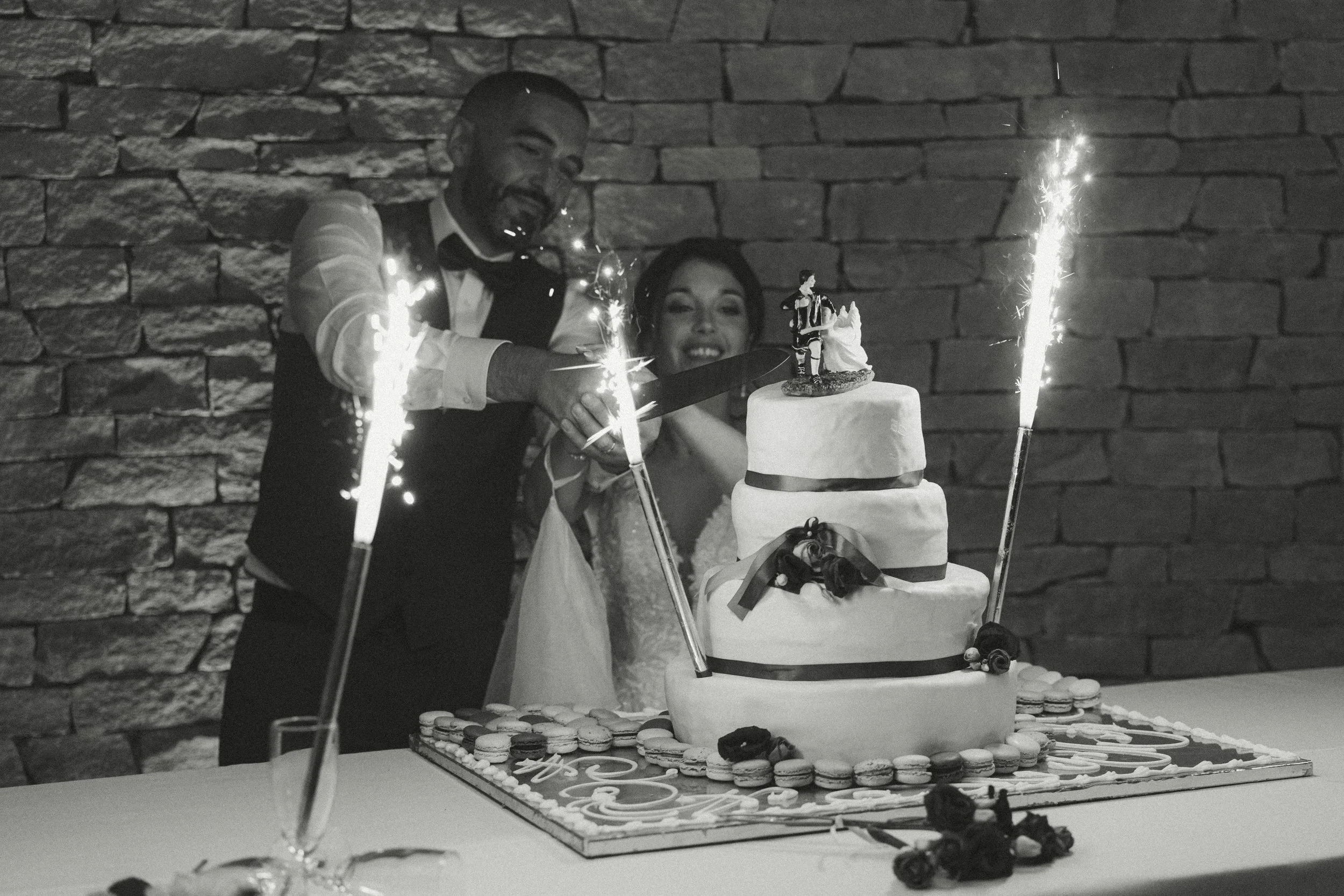 Un couple de mariés souriant coupe un gâteau de mariage à plusieurs étages, entourés de feux d'artifice sur la table.