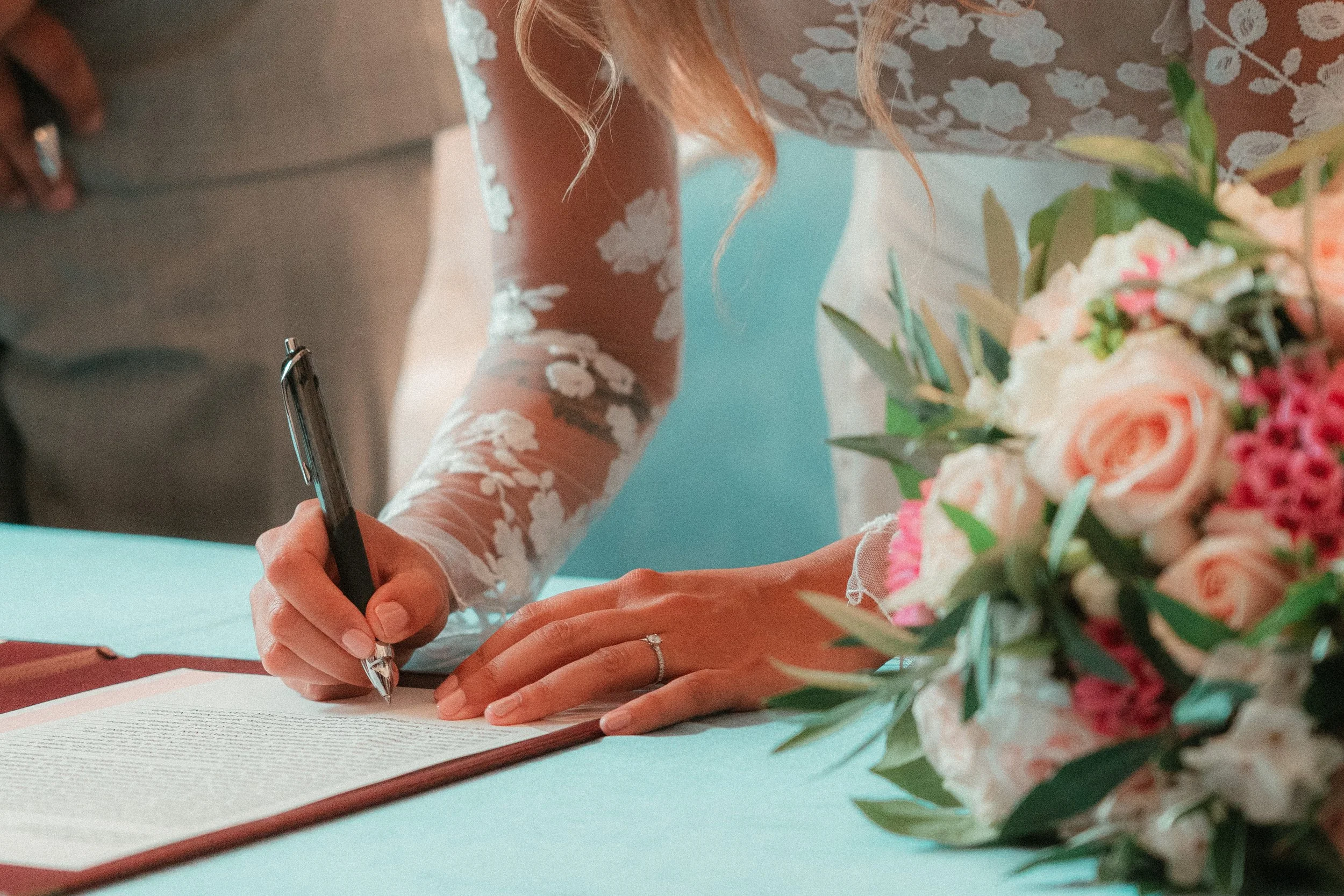 Une femme en robe de mariage signant un document, avec un bouquet de fleurs à côté.