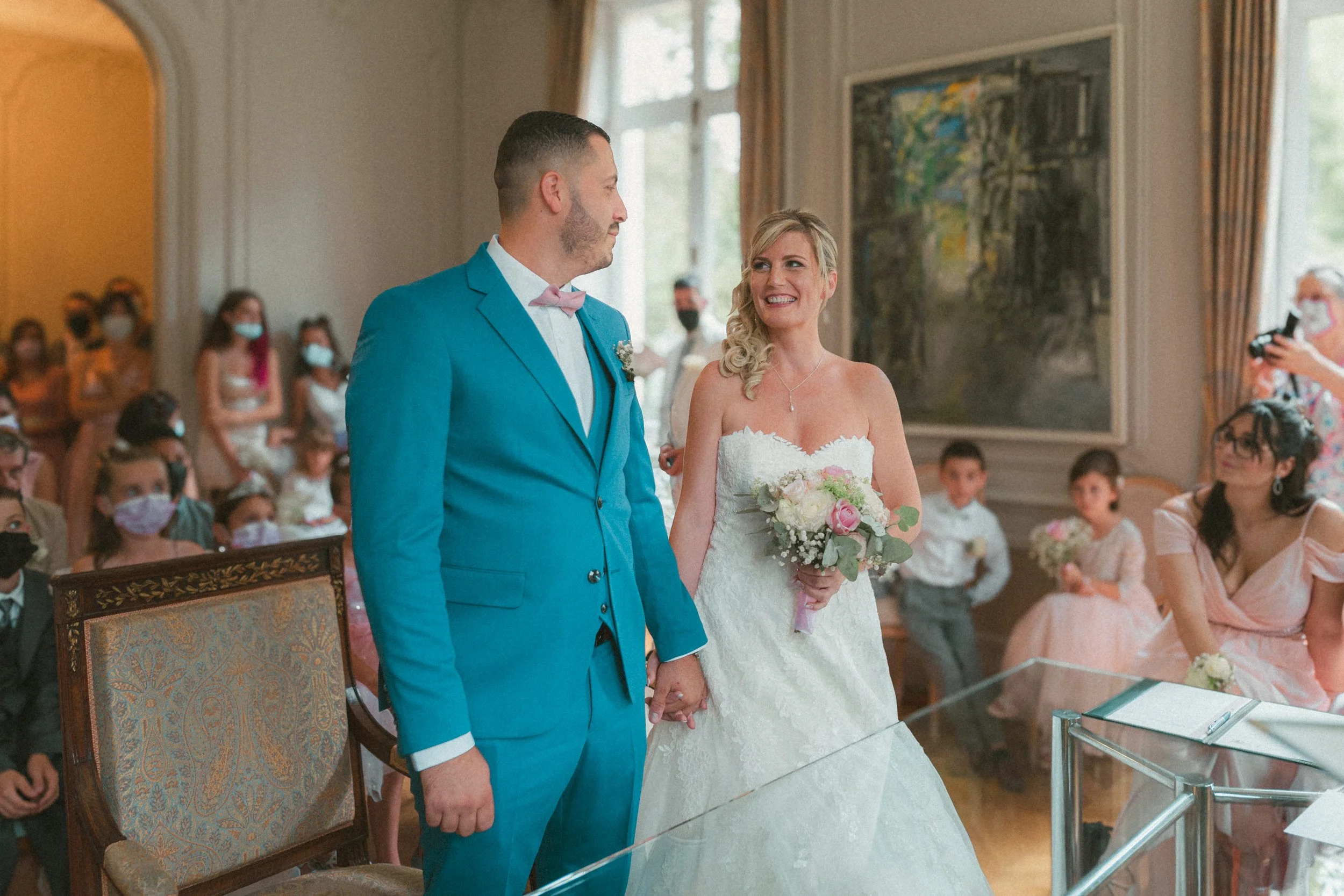 Un couple de mariés tenant la main devant une foule d'assistants à l'intérieur d'une salle de mariage. La mariée porte une robe blanche et tient un bouquet de fleurs, tandis que le marié porte un costume bleu avec une lavallière rose.